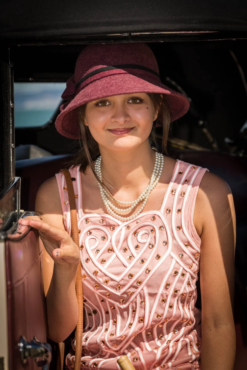 Young woman wearing a pink dress with decorative jewels and a pearl necklace, holding a vintage pink suitcase, and wearing a maroon wide-brim hat, standing inside or near a vehicle.