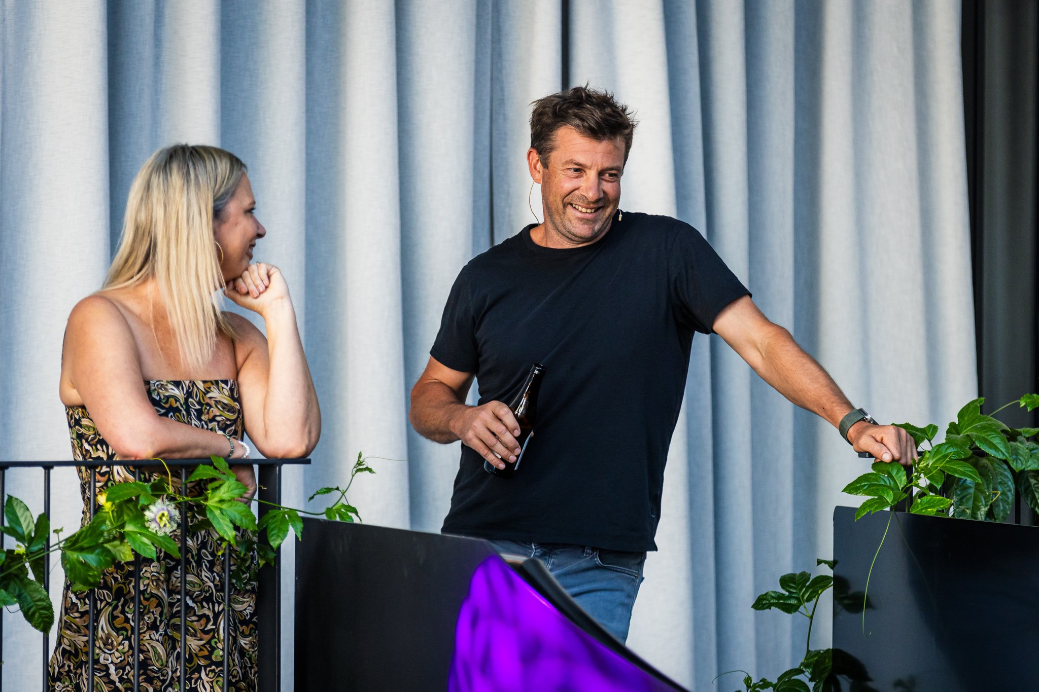 A man and woman standing on a stage with plants, engaged in conversation. The man is smiling, holding a beer, and leaning on a plant pot; the woman is listening, smiling, with her hand on her chin. They are in front of a blue curtain.
