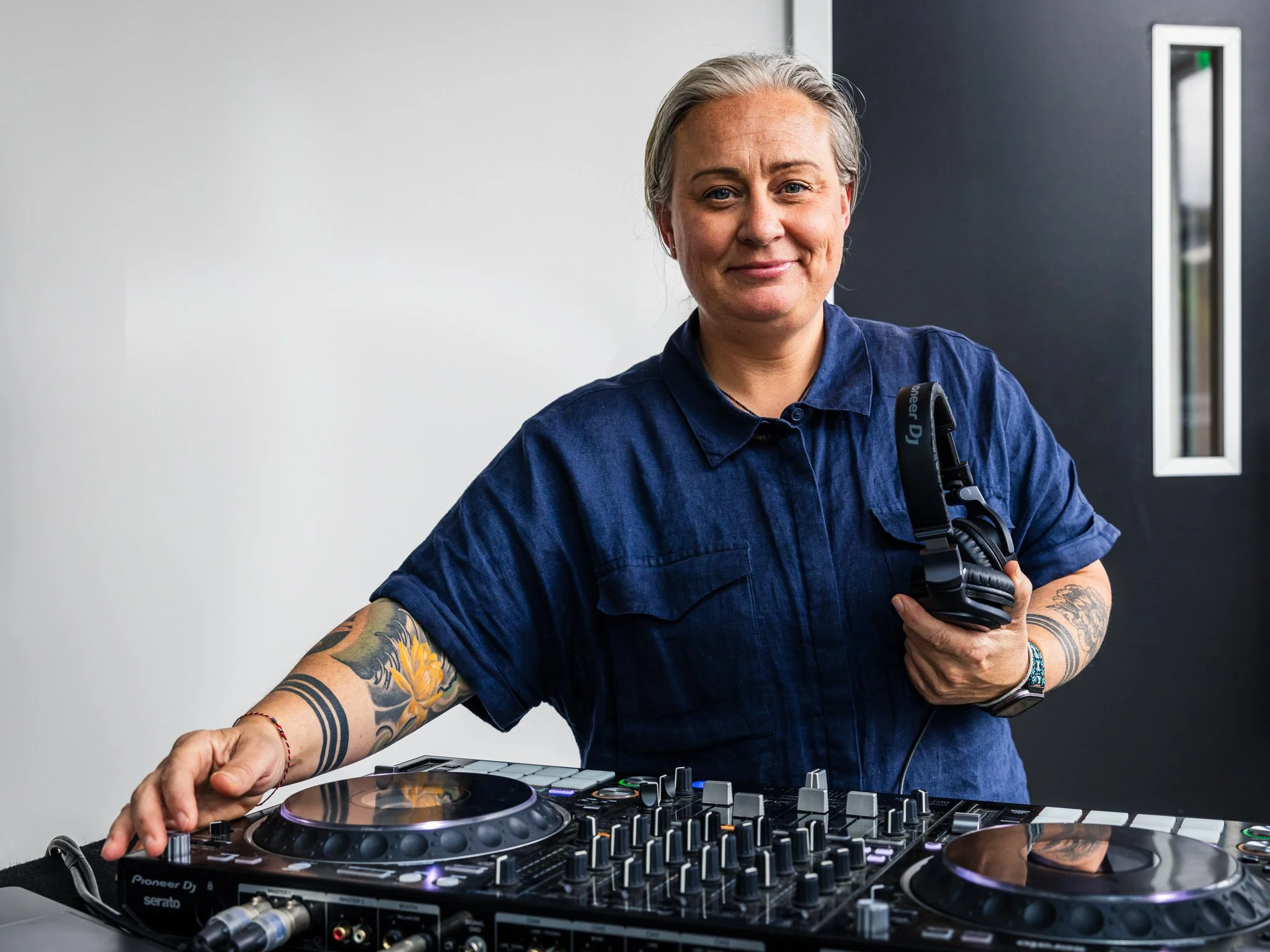 A woman with tattoos on her arms, holding headphones, standing behind DJ equipment in a modern indoor space.