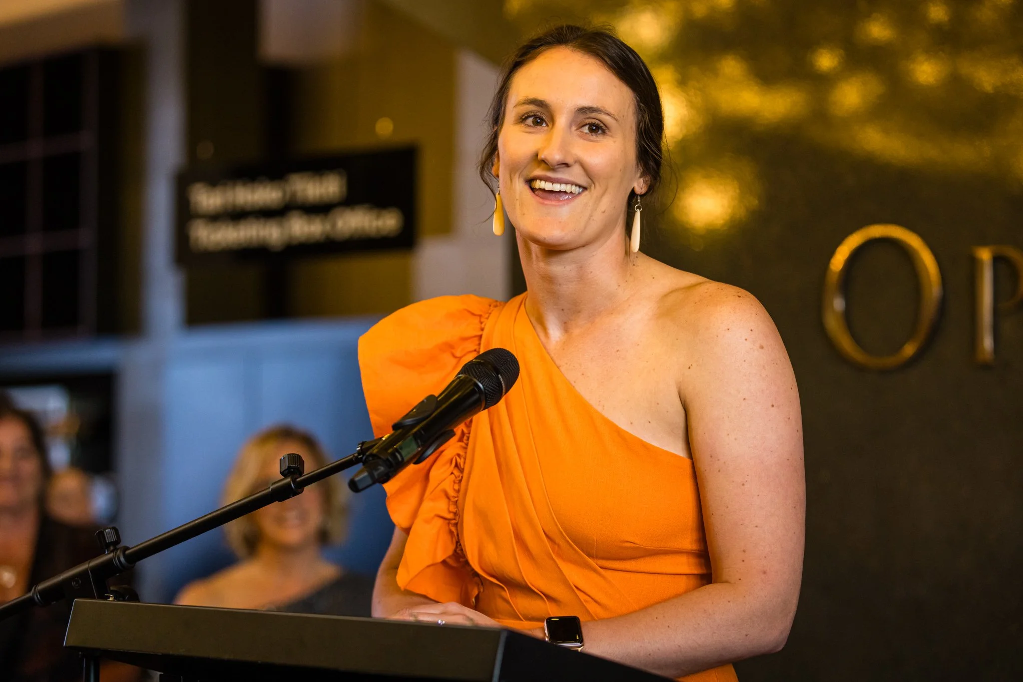 A woman in an orange dress speaking at a podium with a microphone, smiling, with an audience in the background.