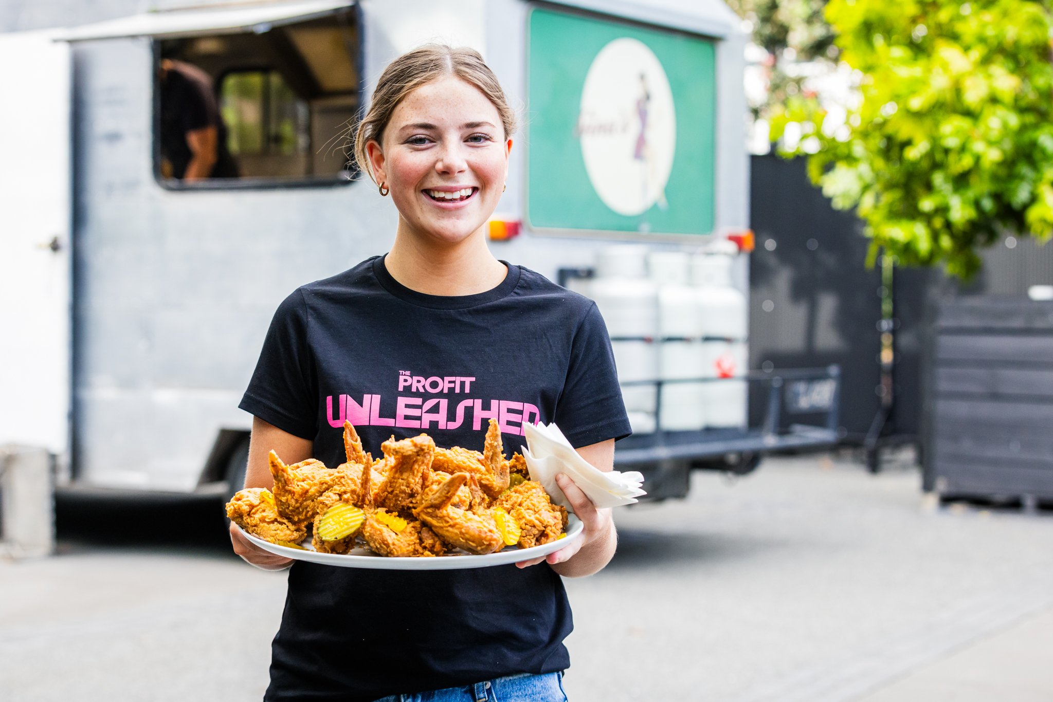 A young woman smiling and holding a plate of fried chicken with pickles and napkins, standing outdoors near food trucks and trees.
