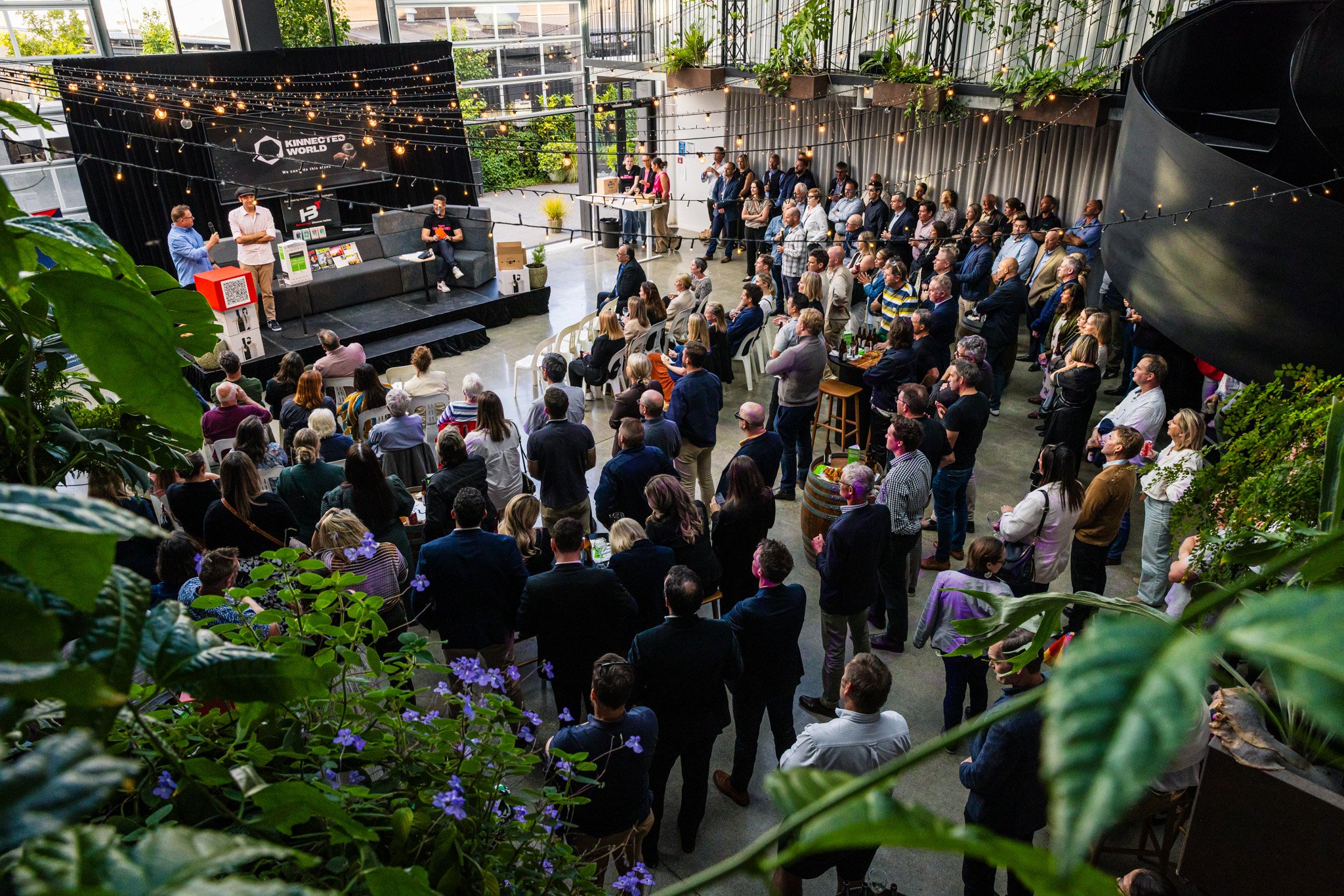 A large indoor event with a stage at the front and many attendees facing it. The stage has a screen with the logo ‘Kinnected World’ and displays speakers, some seated and some standing. The room is decorated with string lights and plants, with some p