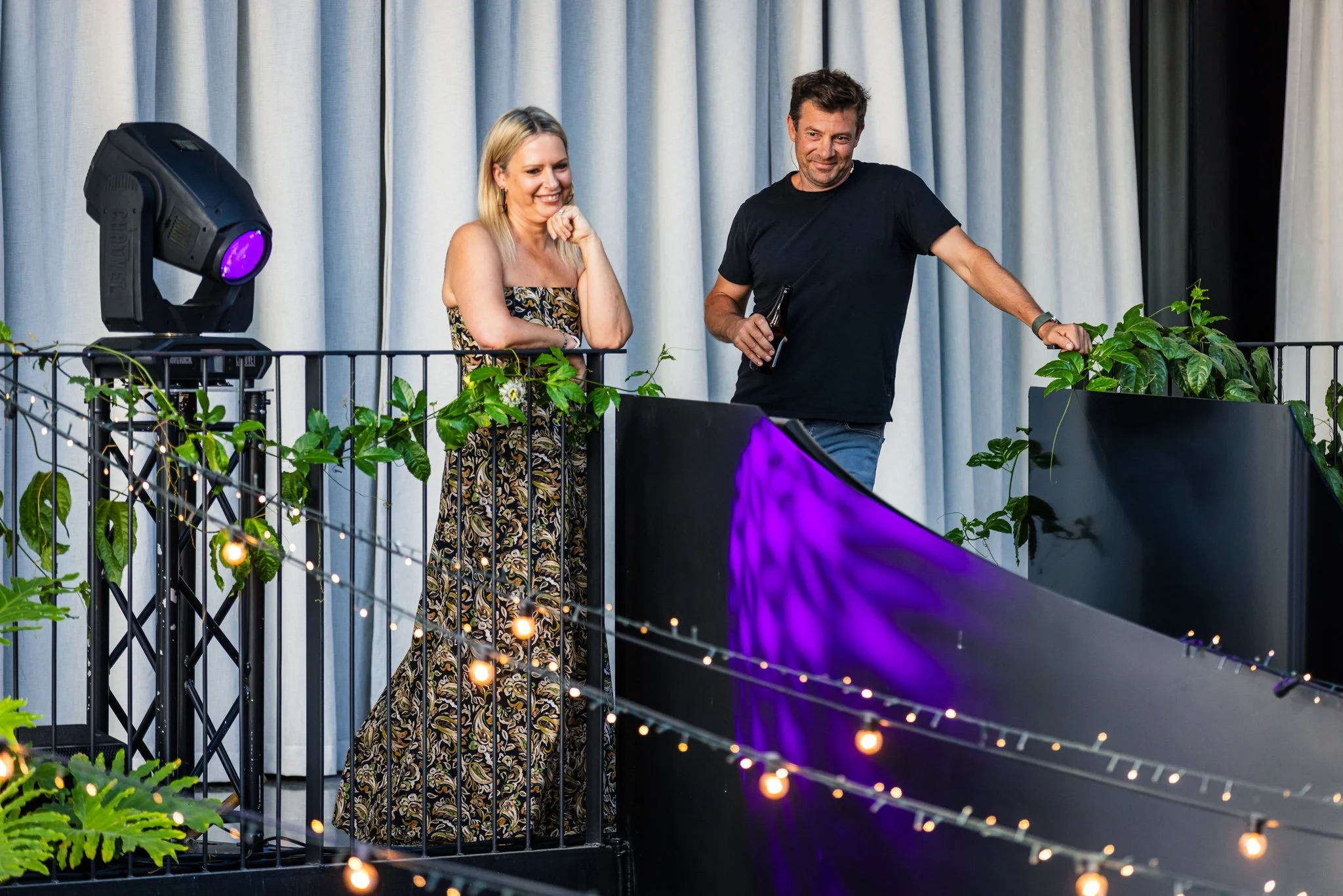 Two people standing on a balcony with plants and string lights. One woman in a strapless patterned dress, one man in a black t-shirt holding a drink, with a large speaker nearby.