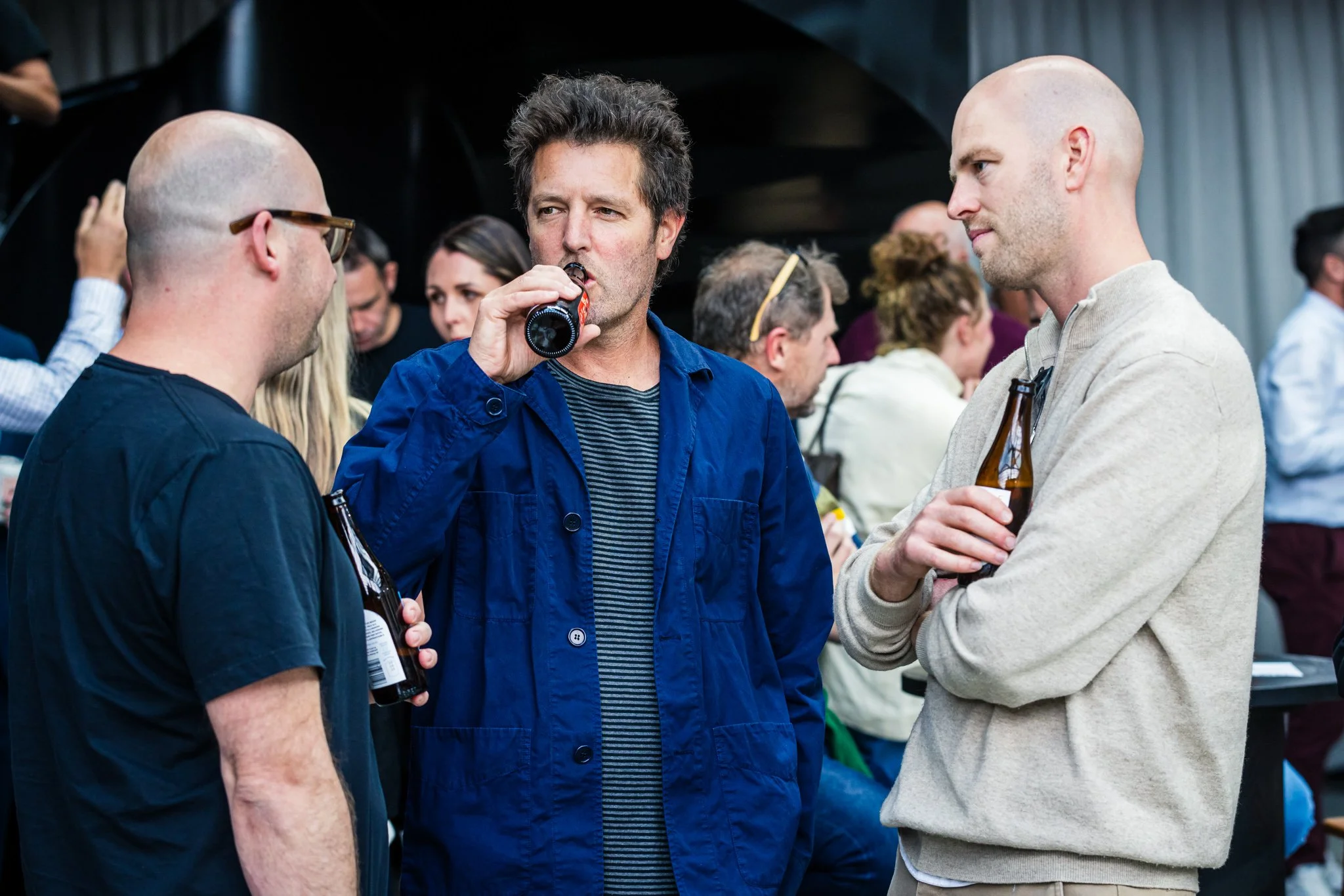 Three men talking and drinking beer at a social gathering, with a group of people in the background.