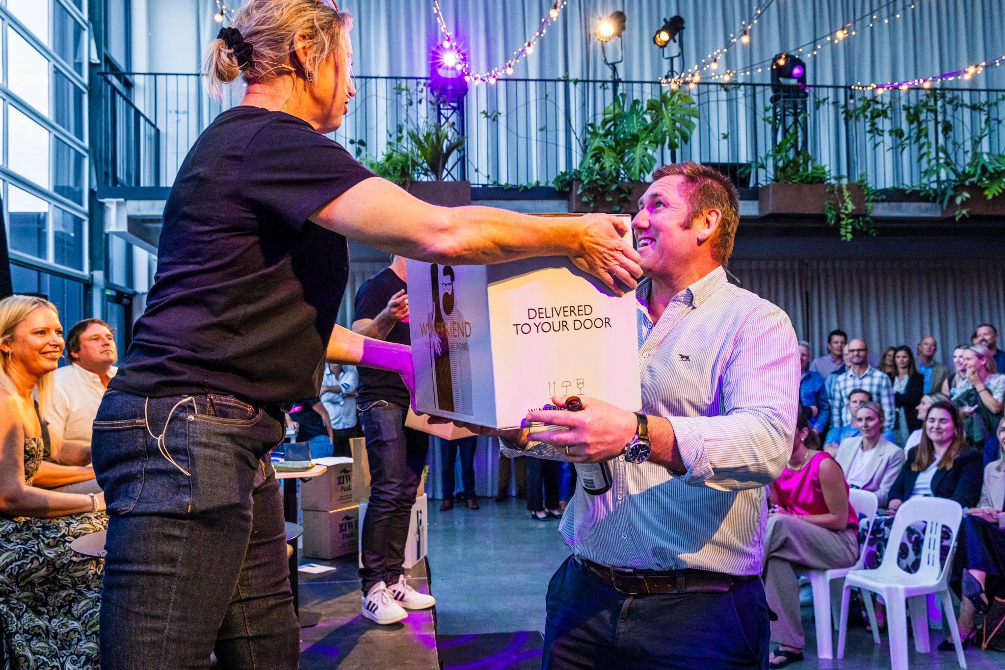 A woman handing a box to a man during an event, with an audience in the background. The box says 'Delivered to Your Door' and has icons of a wine glass, an umbrella, and a wine bottle.