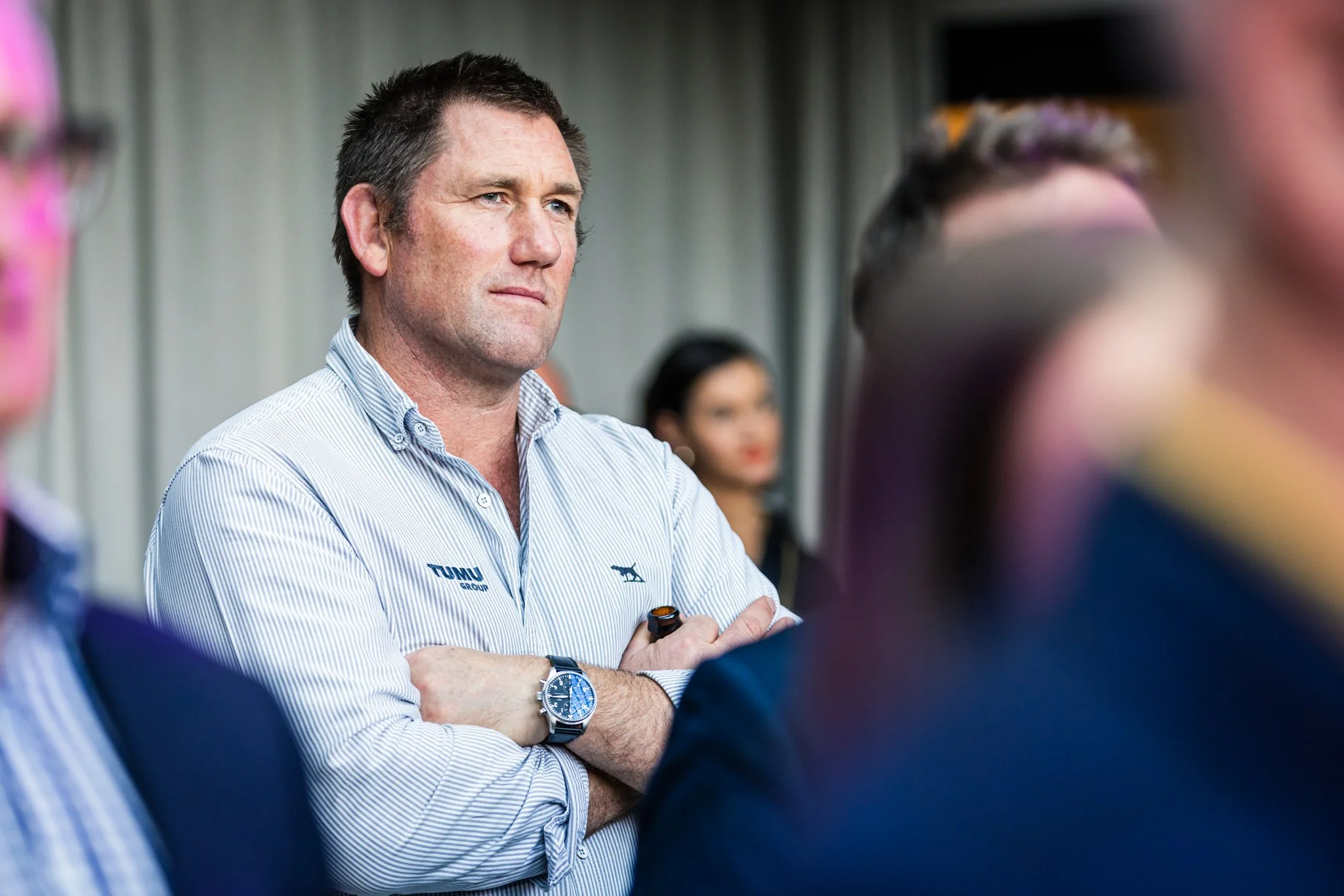 A man with short dark hair, wearing a white and blue striped shirt with the TUMU Group logo, has his arms crossed and is attentively listening during a meeting or event.