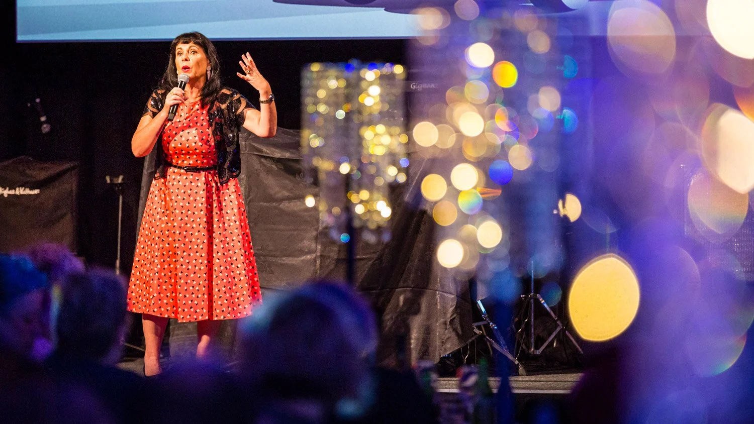 A woman in a red and black dress with a black cardigan speaking into a microphone on stage, with an audience visible in the foreground and colorful bokeh lighting effects.