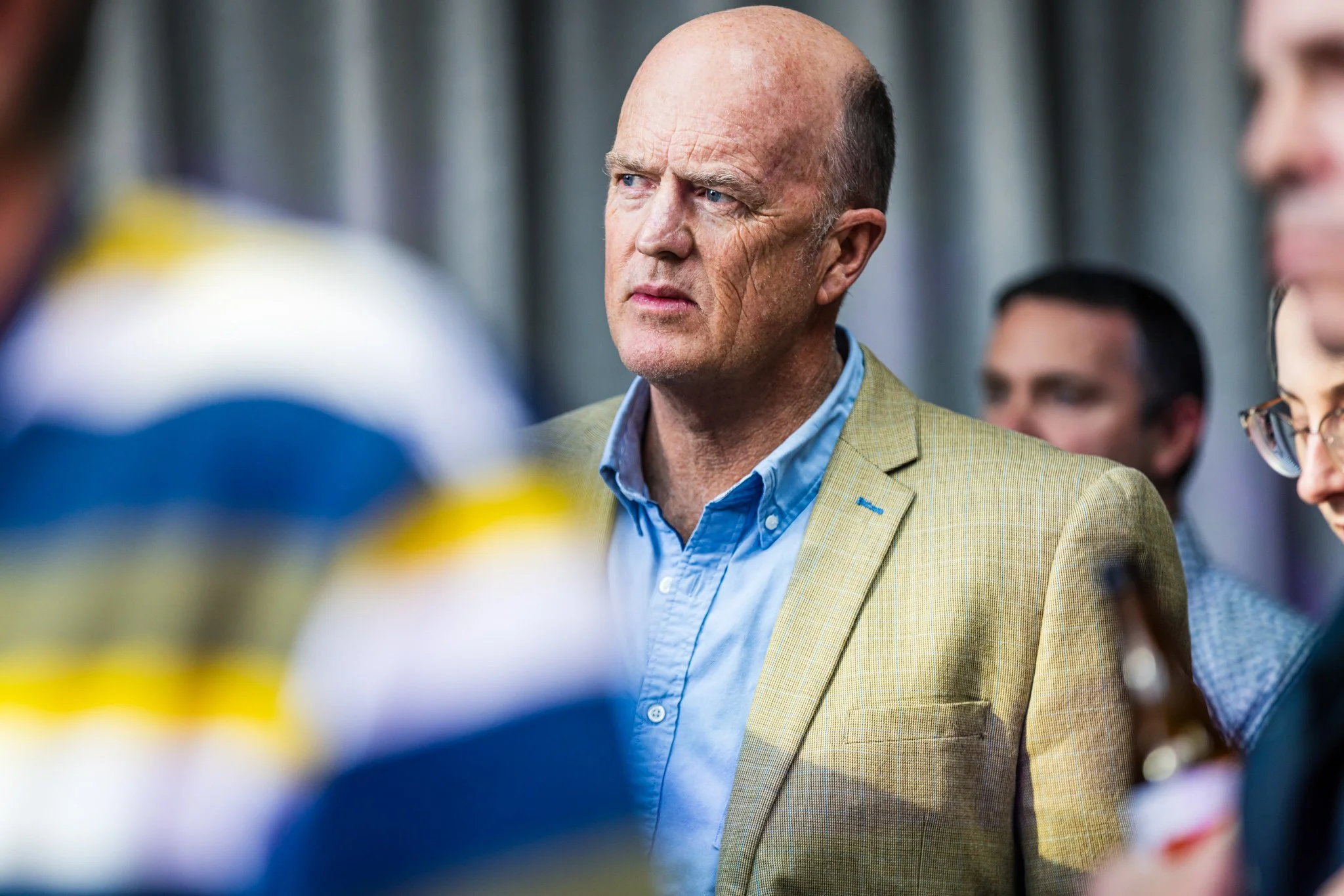 A middle-aged man with a serious expression, wearing a light beige blazer and blue shirt, standing among a group of people engaged in conversation.