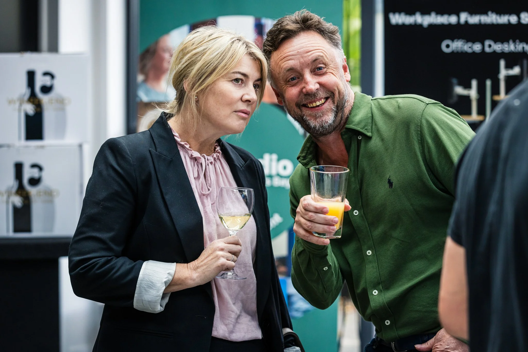 Two adults, a woman and a man, at a social event holding drinks and engaging in conversation with a person whose back is to the camera.
