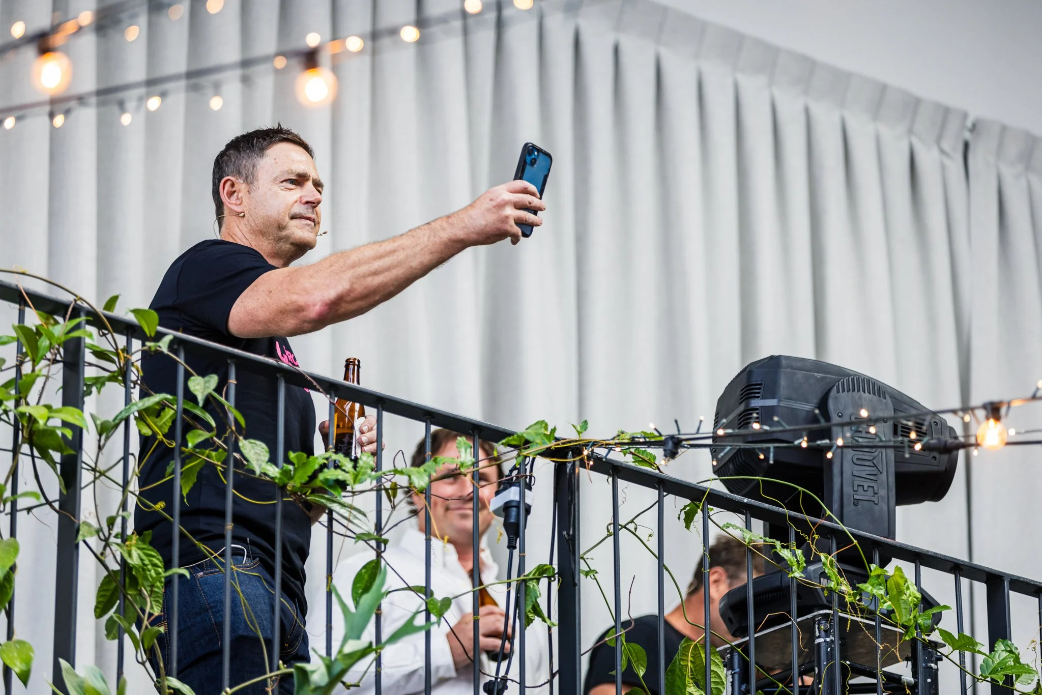A man is taking a selfie with his phone at a social gathering. He is standing on a balcony or raised platform, holding a Beer bottle in his other hand. There are string lights hanging above, and some greenery is visible around the railing. In the bac