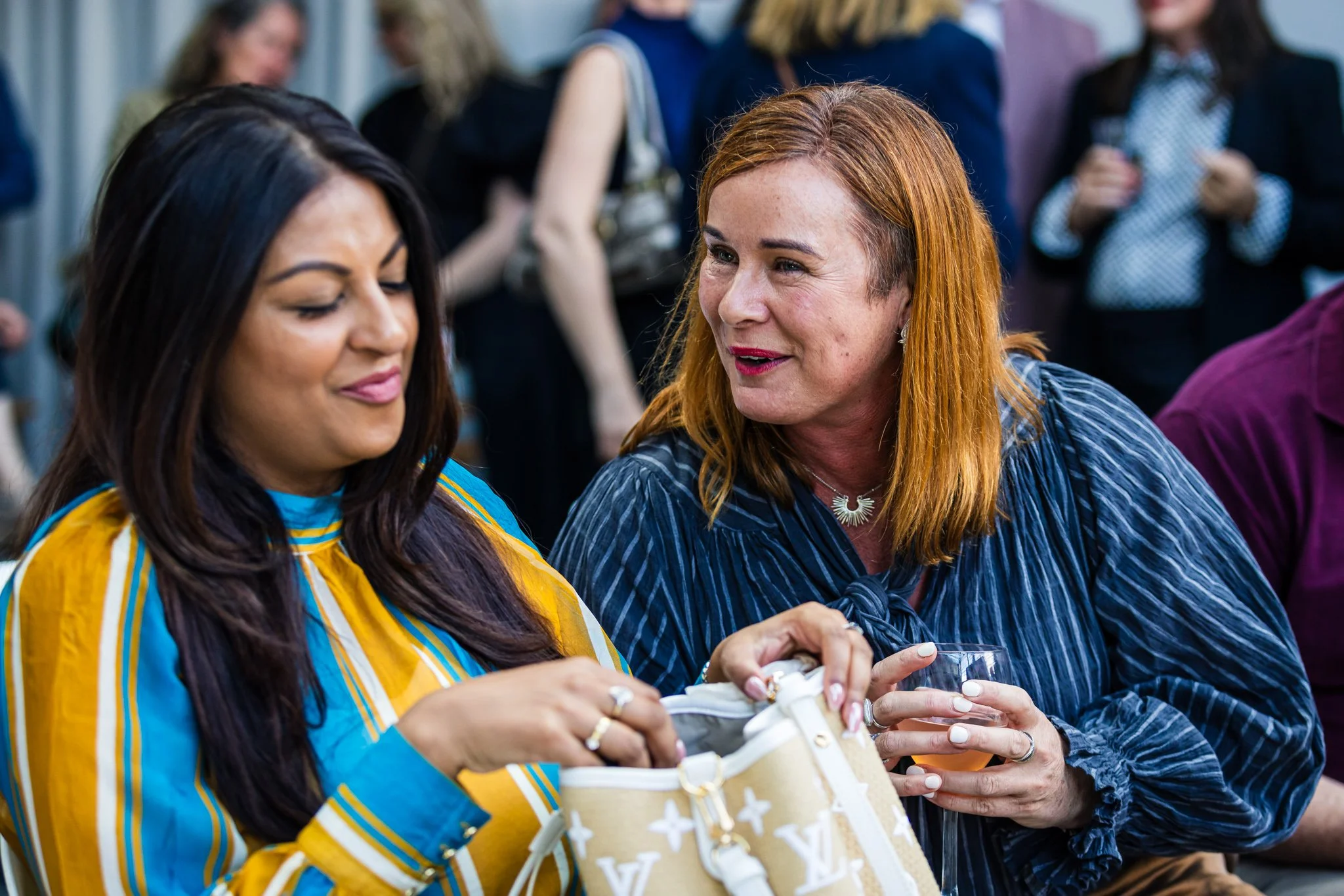 Two women at a social event, one with dark hair wearing a yellow and blue striped shirt, and the other with red hair and a blue striped blouse, engaged in conversation while holding a drink and a small purse.