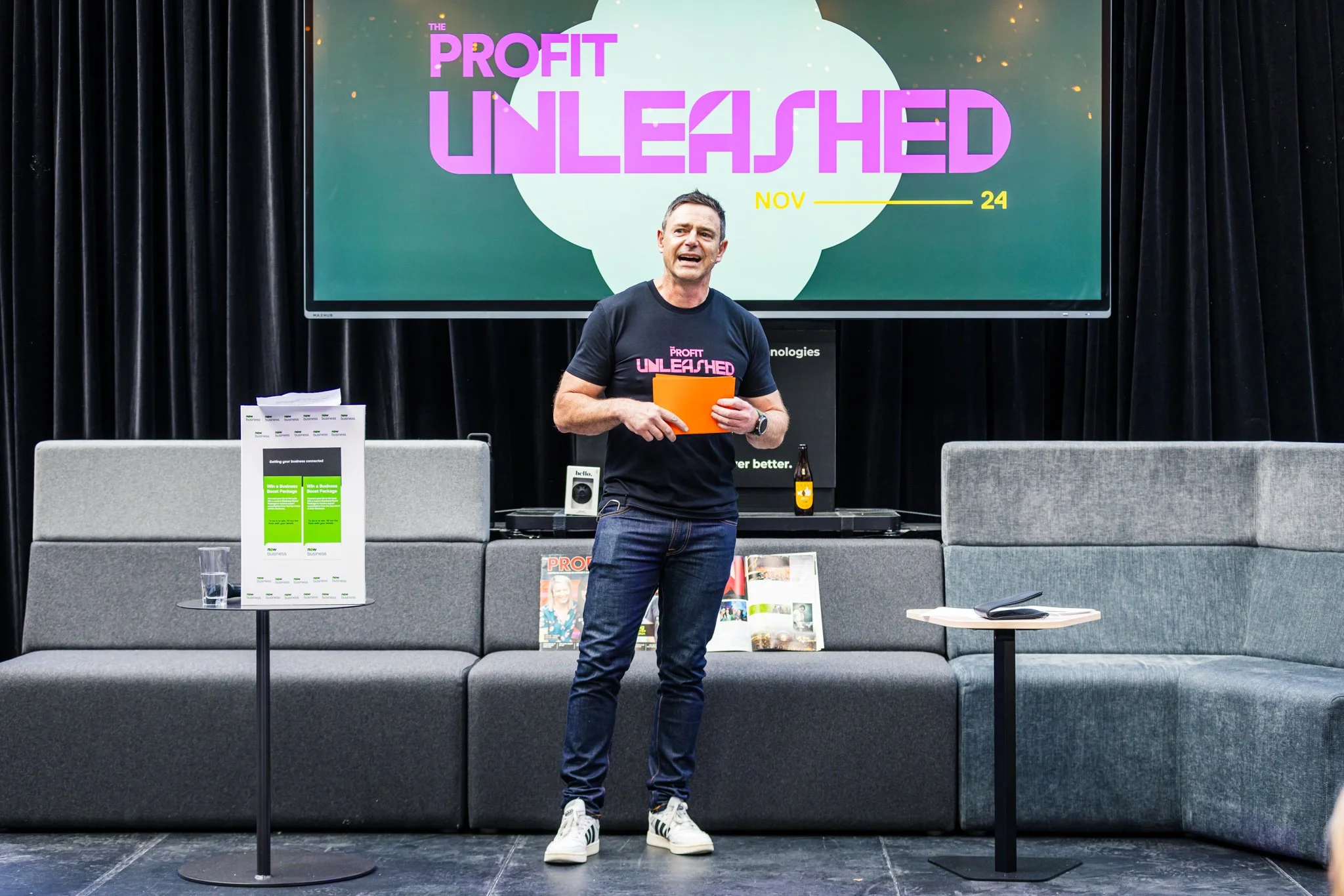 A man wearing a black T-shirt with 'Profit Unleashed' written on it, standing on a stage with a microphone clipped to his shirt, holding an orange folder. Behind him is a large screen displaying 'Profit Unleashed' and the date 'November 24'. The stag