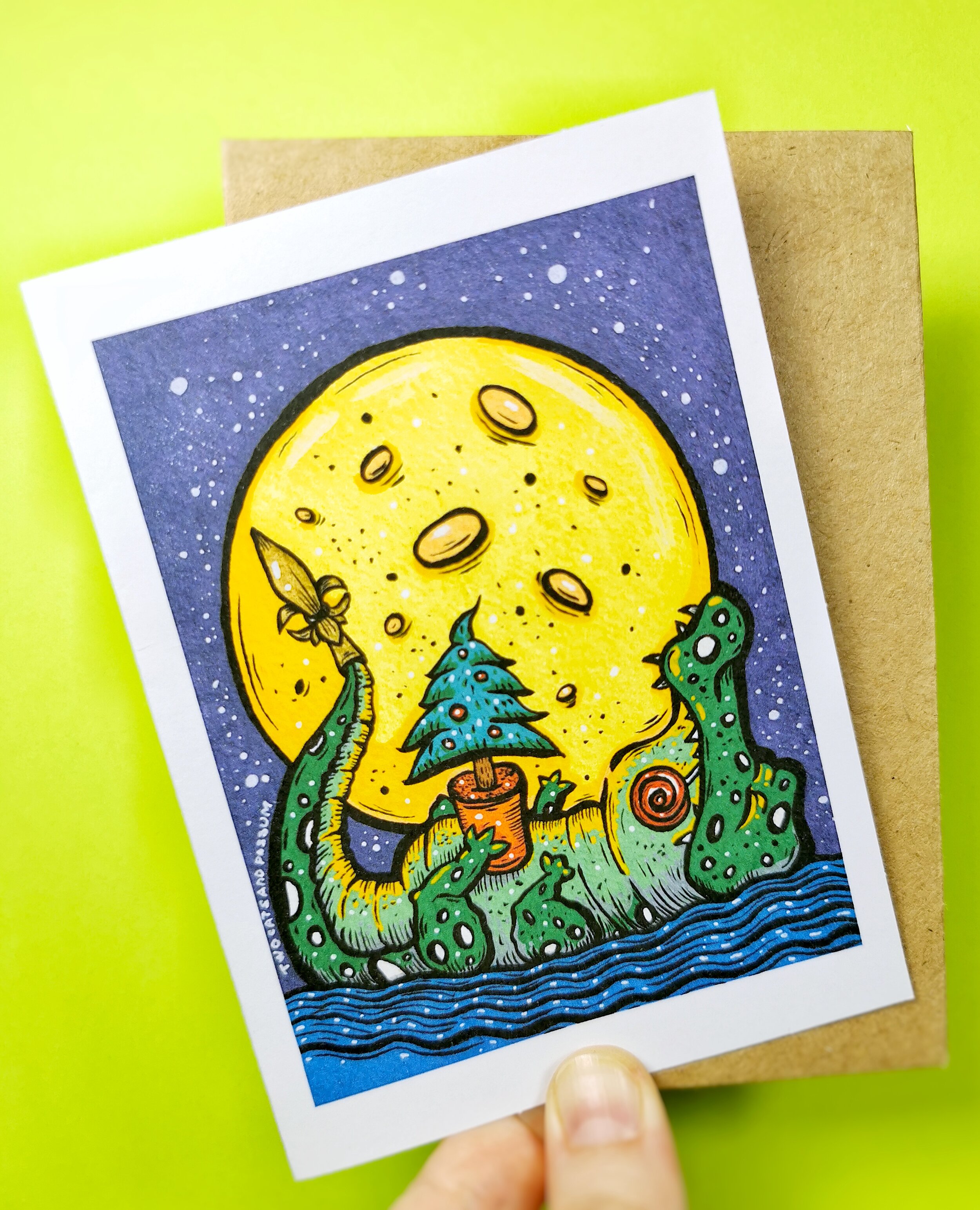fingers holding Christmas card featuring alligator with Christmas tree and full moon