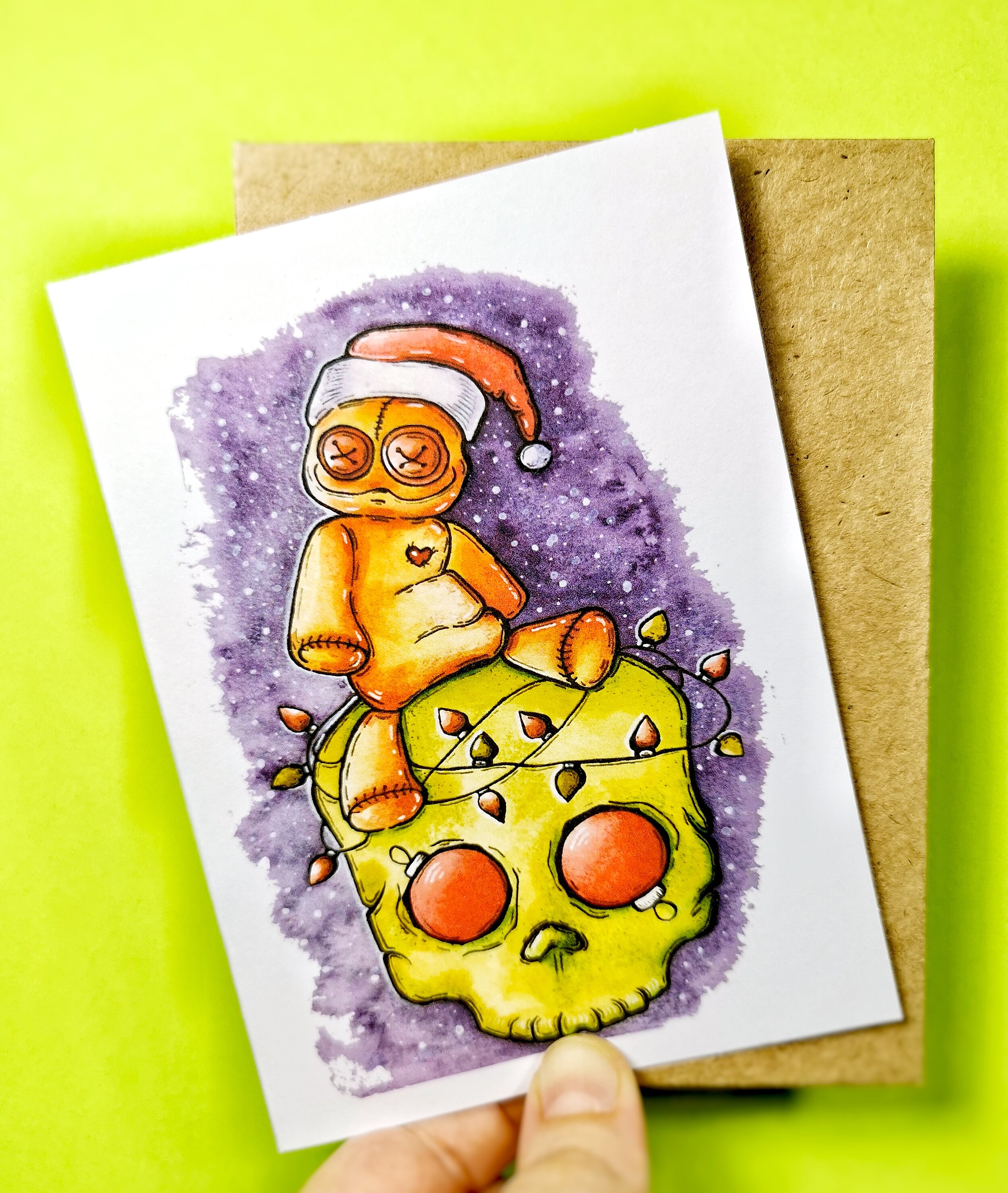 fingers holding Christmas card featuring voodoo doll sitting on skull with Christmas lights and ornaments