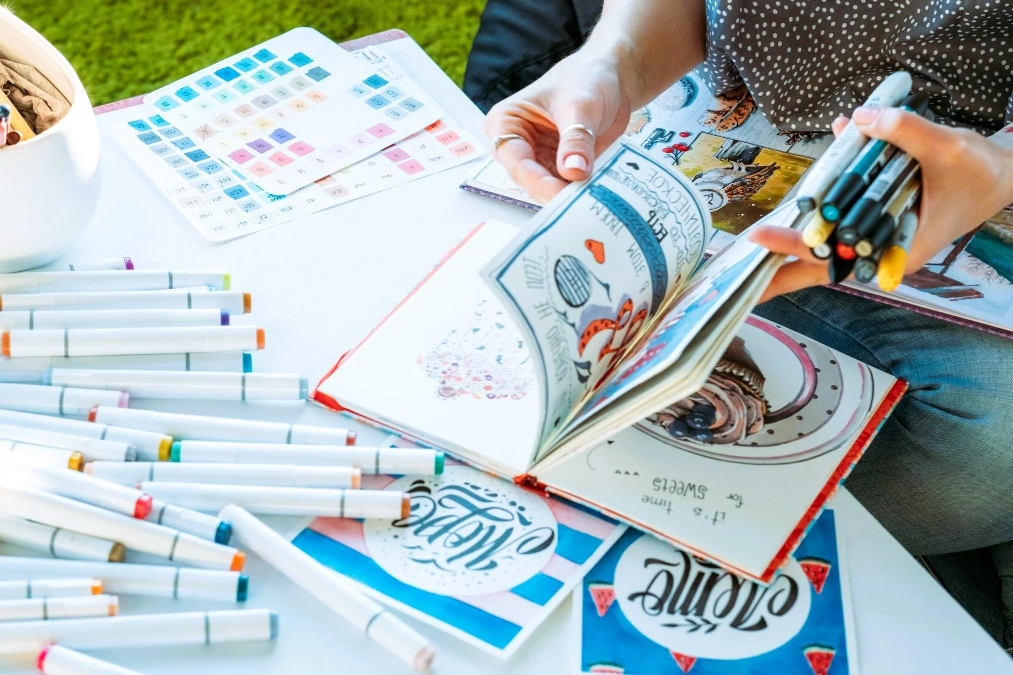 An artist surrounded by alcohol markers, color swatches, sketchbook and illustrations