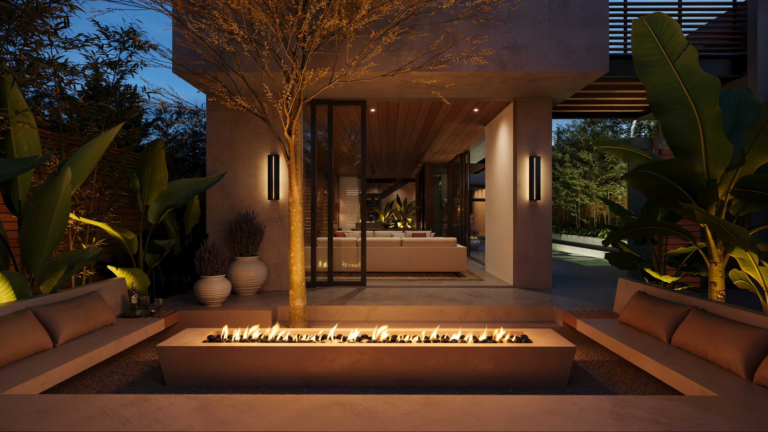 Modern outdoor patio with a central linear fire feature, surrounded by cushioned lounge seating, potted plants, trees, and ambient lighting, overlooking a stylish home interior at dusk.