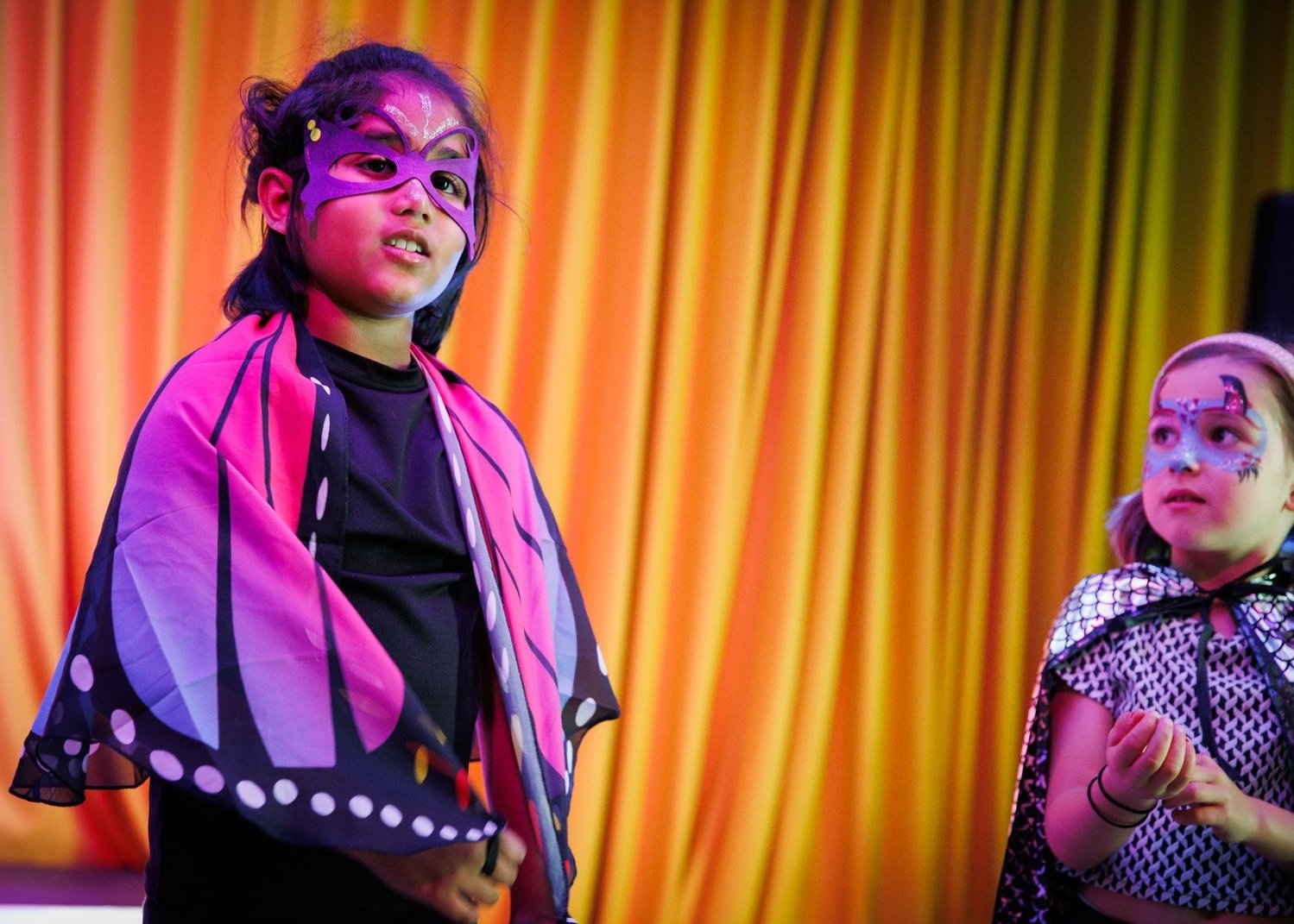 BLAST Youth Theatre members: Children wearing colorful butterfly and superhero costumes with face paint, standing in front of a yellow curtain.