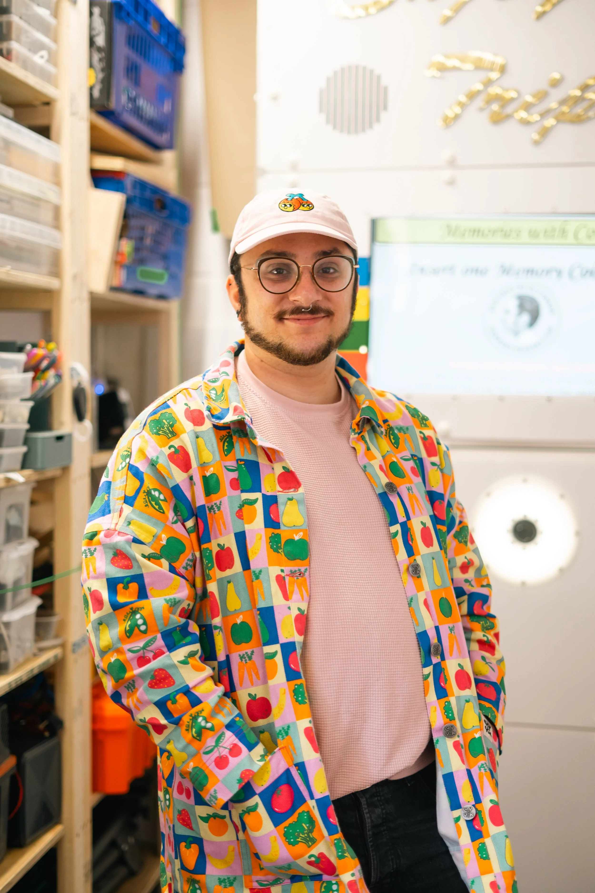 Person wearing colorful fruit-patterned shirt and light pink cap, standing in a room with shelves and bins in the background.