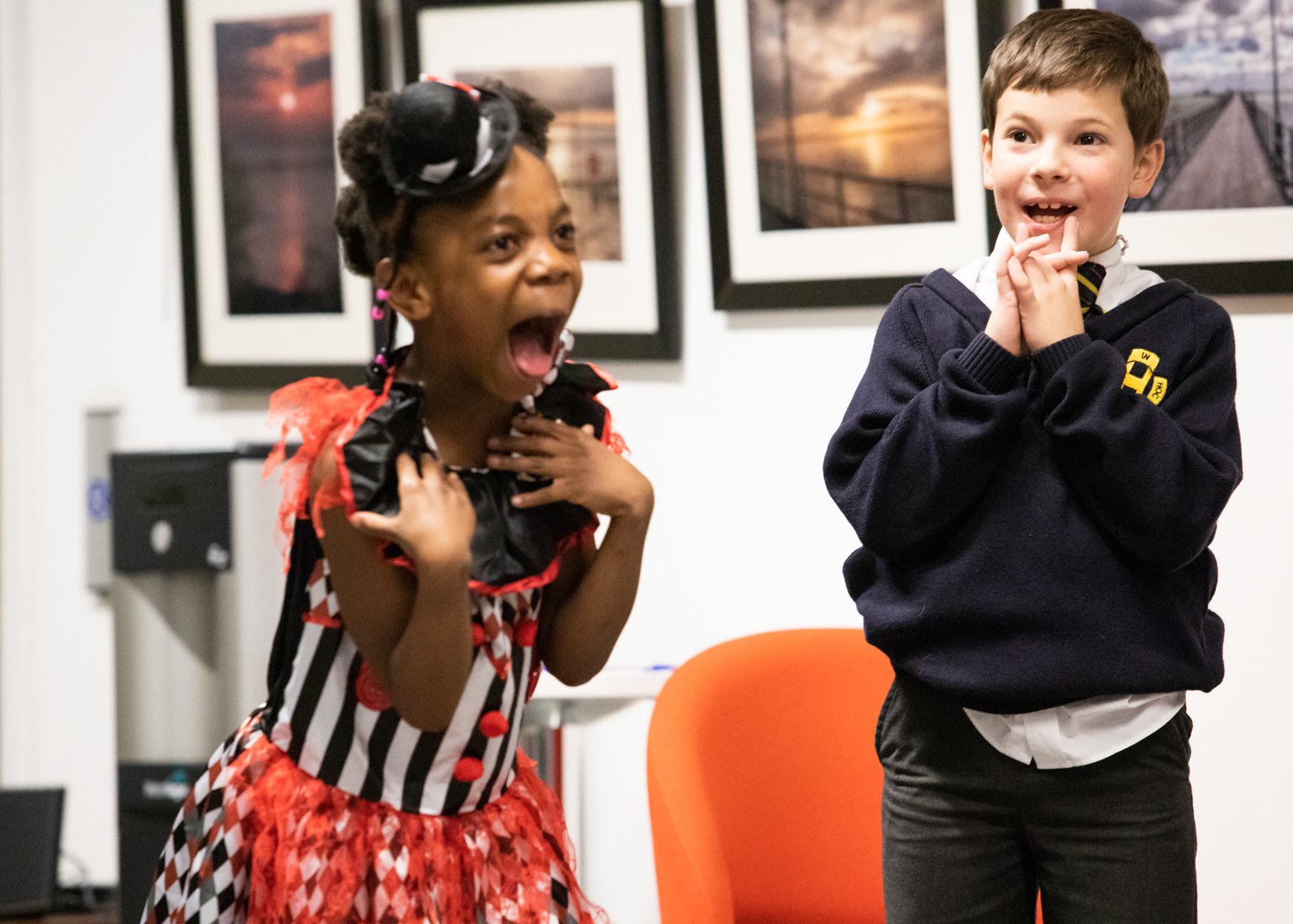 A young girl dressed as a clown and a young boy in a school uniform are standing indoors, both showing surprised or excited expressions.