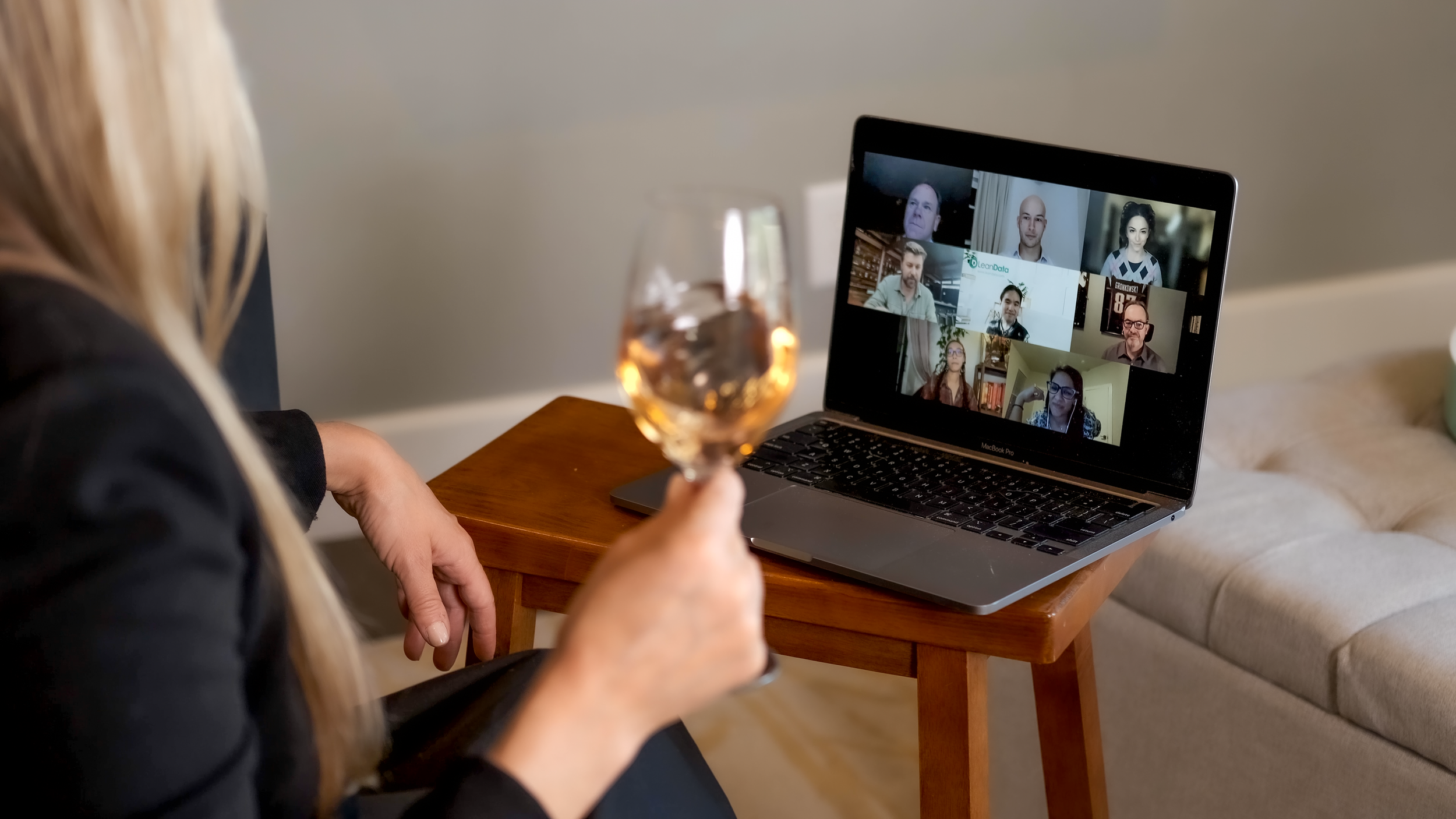 Woman holding a glass of rosé wine while participating in a virtual meeting on a MacBook Pro, with multiple participants visible on the screen, sitting at a wooden table in a cozy room.