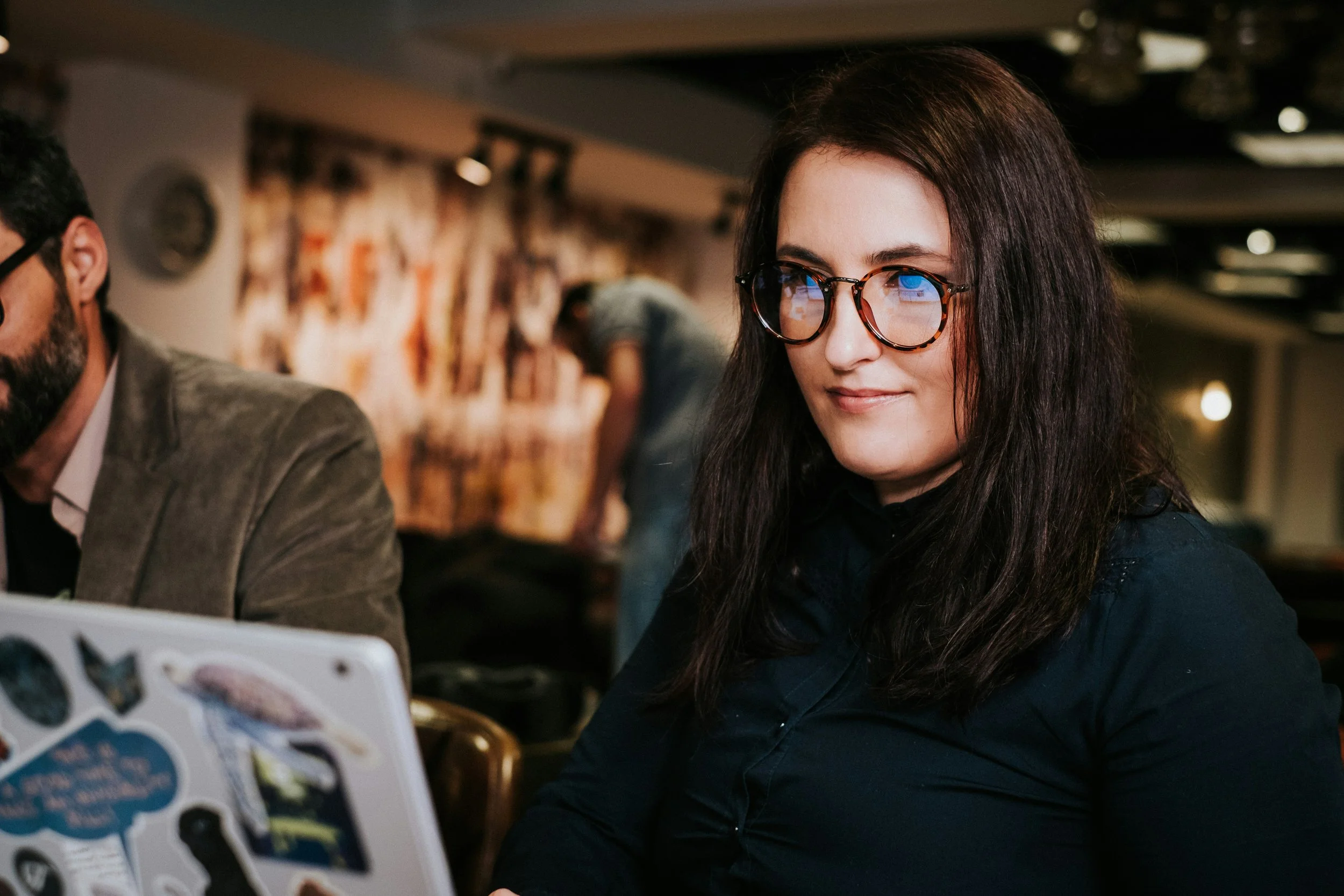 A woman with long dark hair wearing glasses and a dark top, sitting indoors, smiling and looking slightly to her right.
