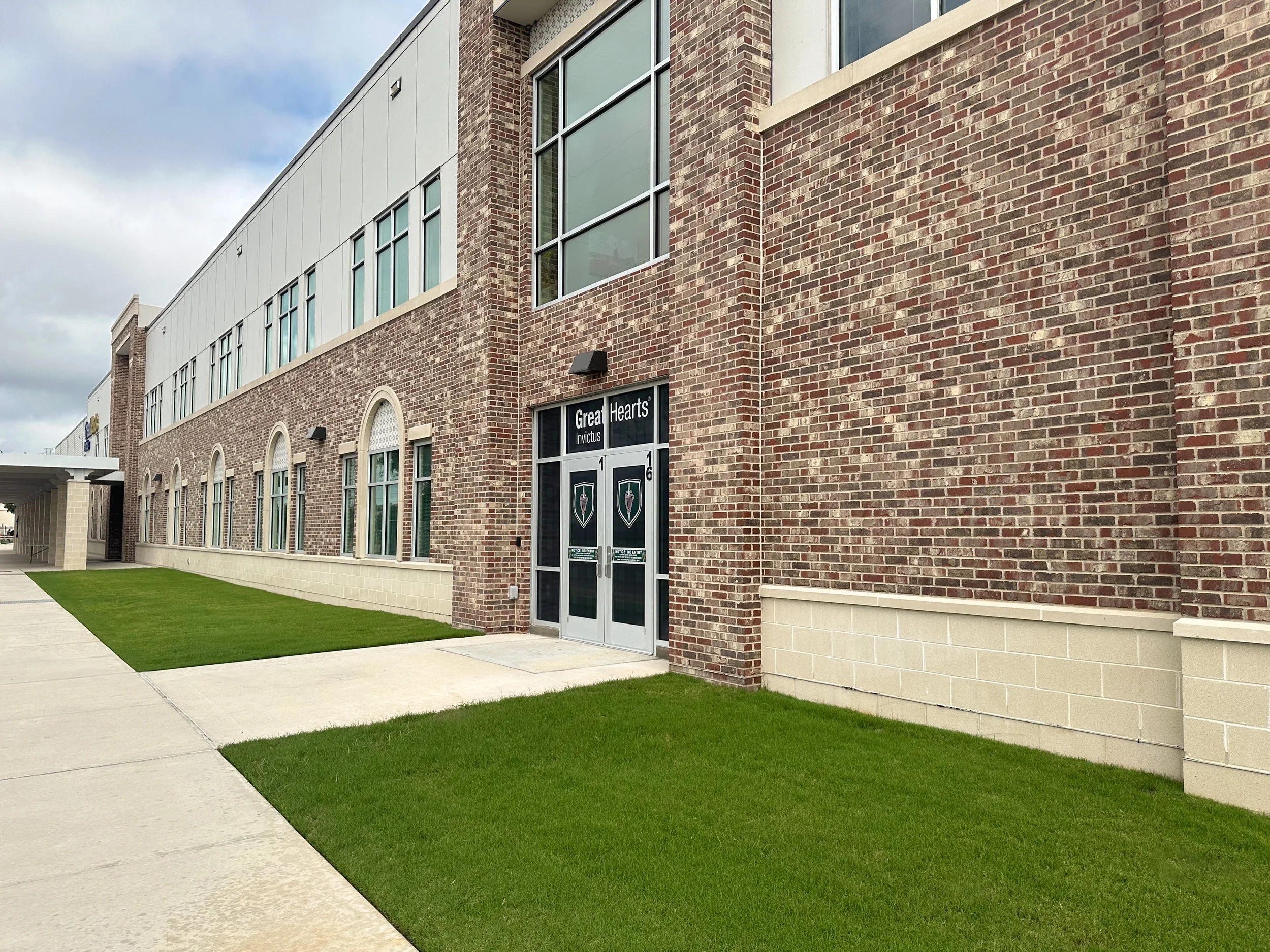 Exterior of a brick building with large windows, a sign that says 'Great Hearts,' and a grassy area with a sidewalk.