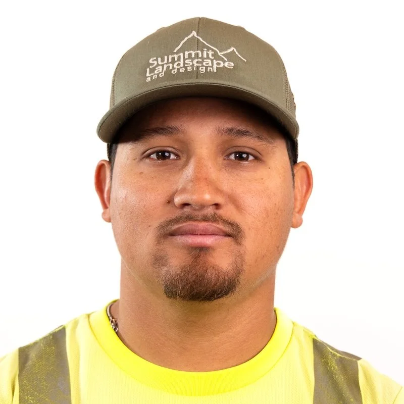 A man wearing a tan baseball cap with white text and mountain outline, a yellow safety vest, and a chain necklace, against a white background.
