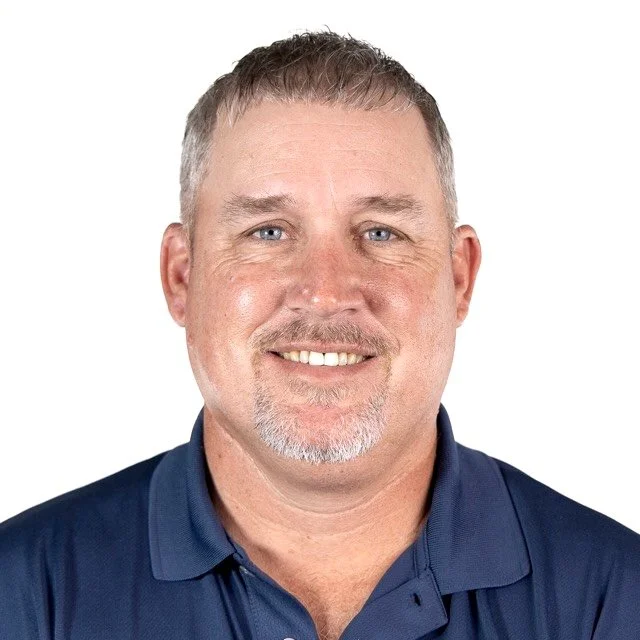 Close-up of a middle-aged man with short gray hair, a goatee, blue eyes, and a friendly smile, wearing a navy blue polo shirt against a white background.