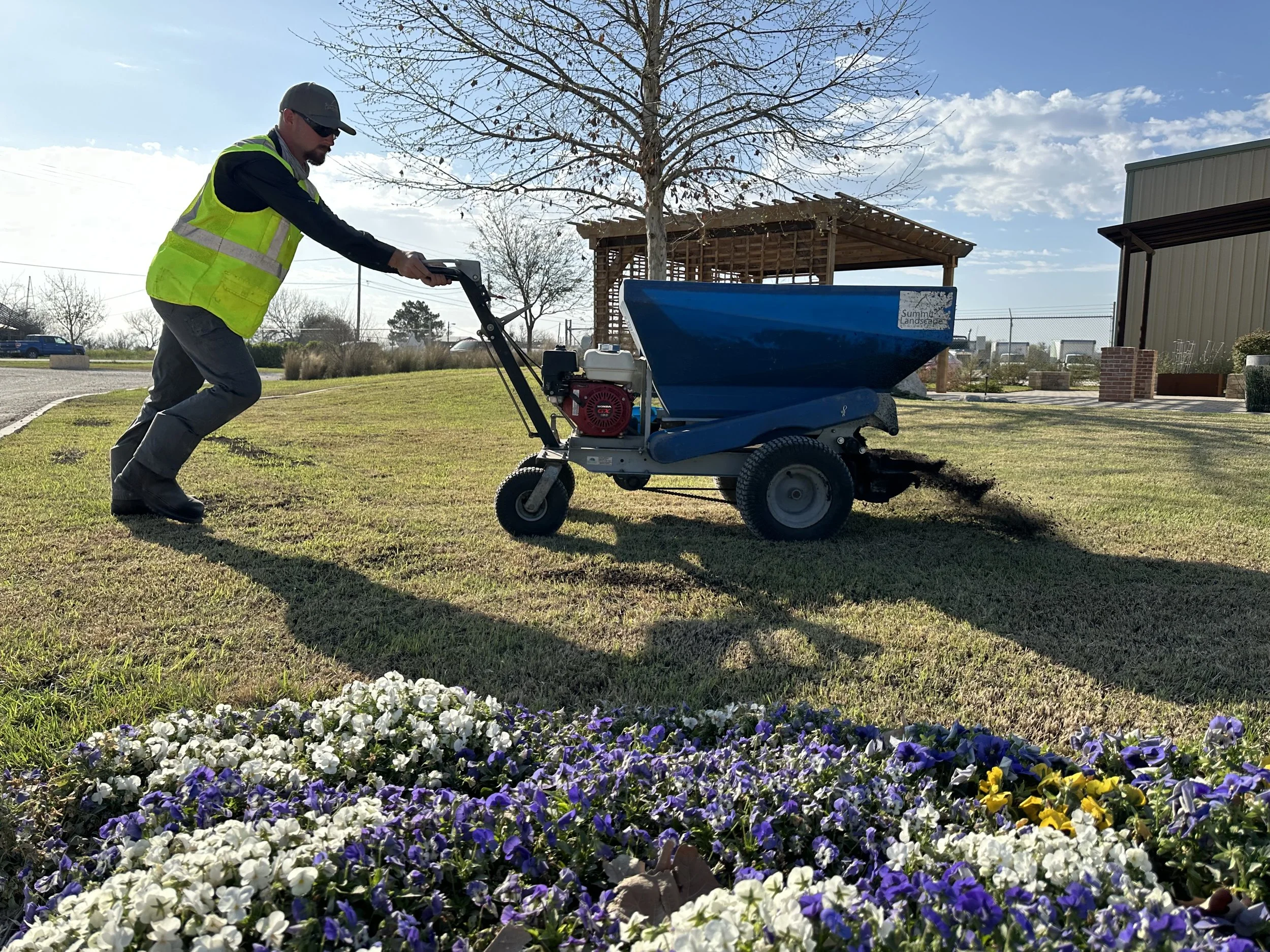 A man in a yellow safety vest using a power machine to level the soil on a lawn near purple, white, and yellow flowers.