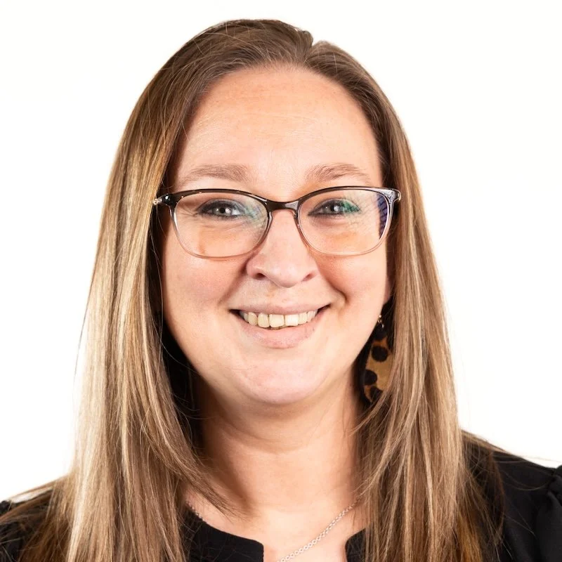 A woman with long straight hair wearing glasses, earrings, and a black top, smiling against a white background.