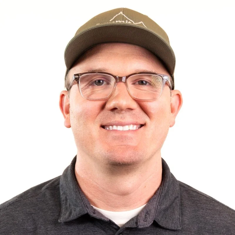 A smiling man wearing glasses, a tan cap, and a dark gray collared shirt against a white background.