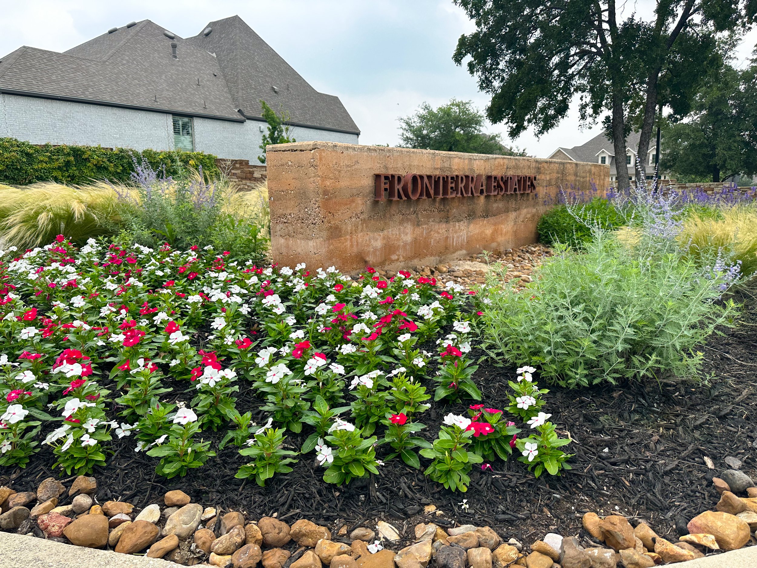 A landscaped garden with pink and white flowers, green plants, and ornamental grass surrounding a brick sign that reads "FRONTERRA ESTATES," with houses and trees in the background.