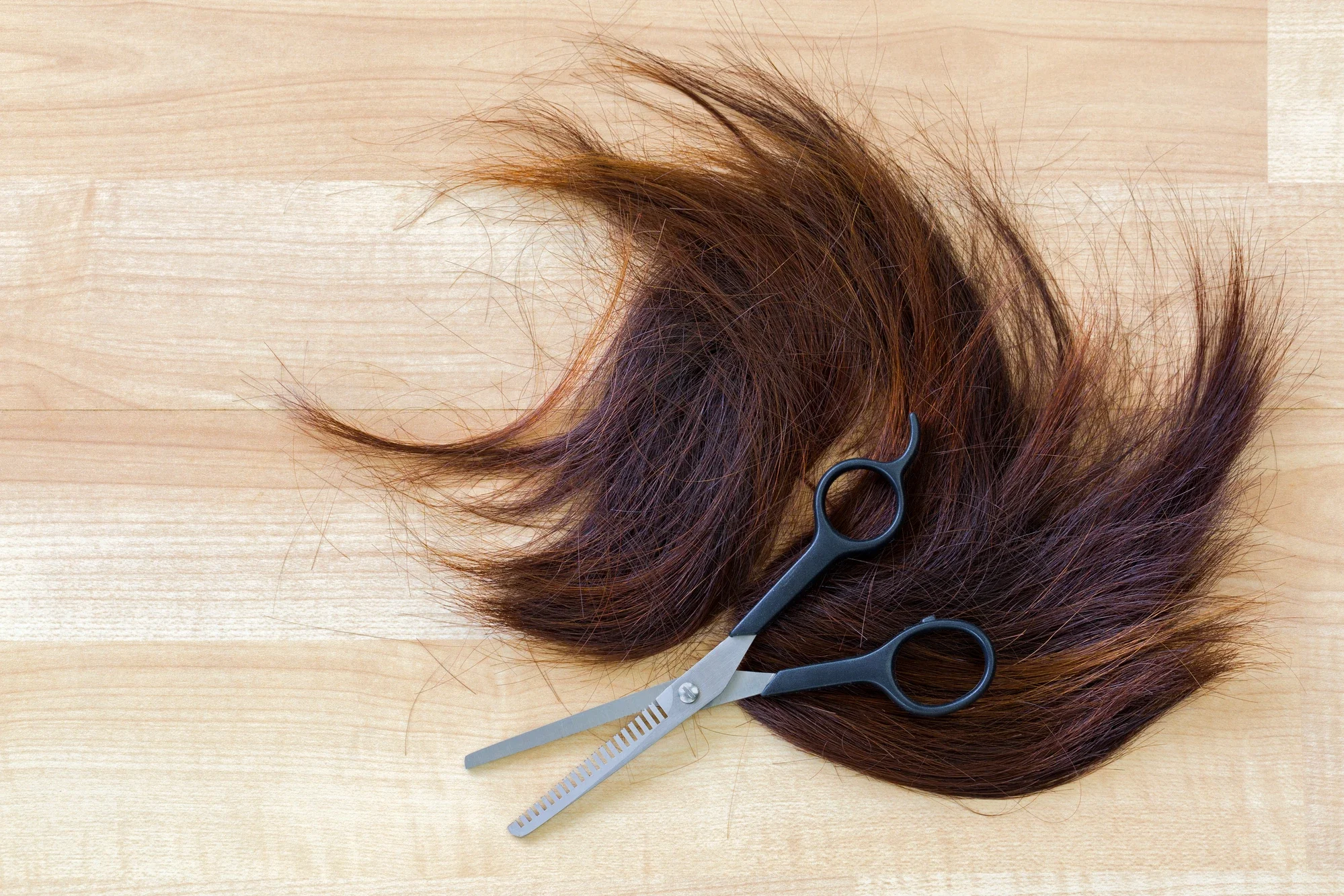 A pile of freshly cut brown hair lies on a light wooden surface beside a pair of thinning shears.