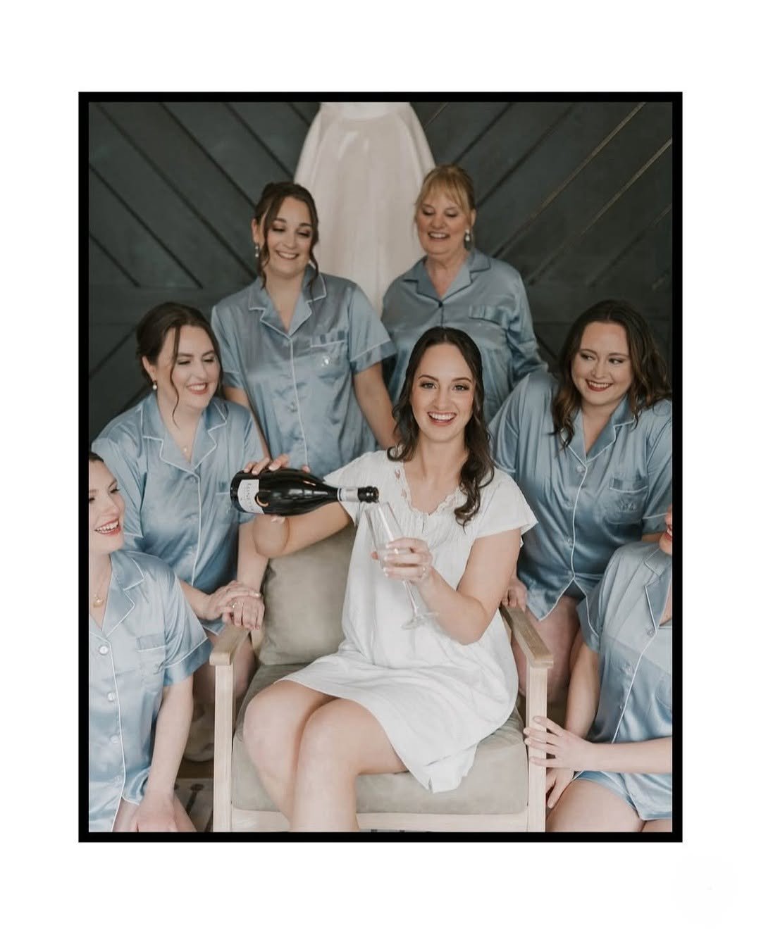 The bride, surrounded by her bridesmaids in matching blue satin pajamas, smiles while pouring champagne—hair and makeup by TEAL Hair and Makeup Studio.