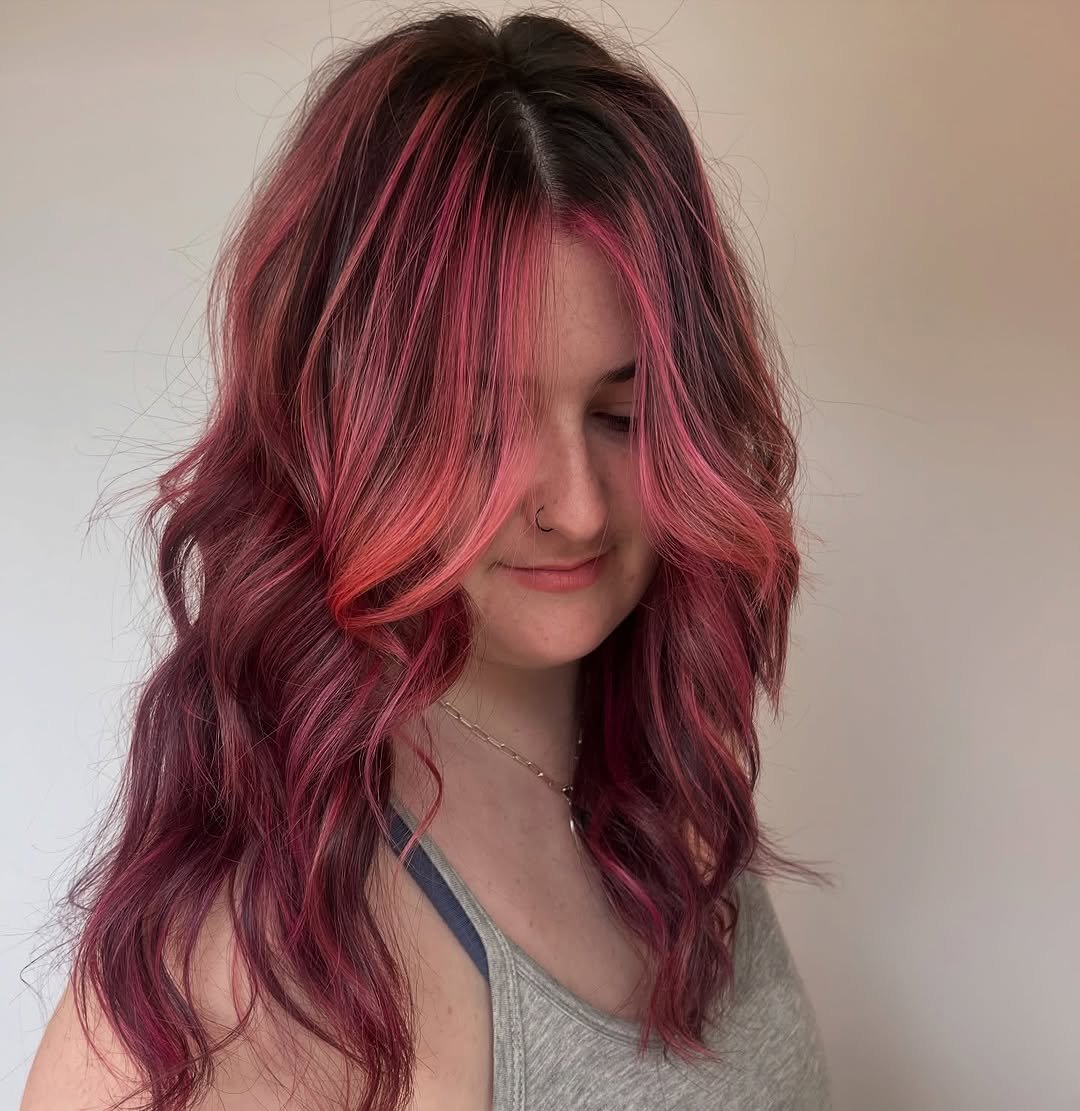 A client at TEAL Hair and Makeup Studio showcases vibrant, wavy hair with rich berry and hot pink highlights, styled in voluminous loose curls and a soft face-framing finish.