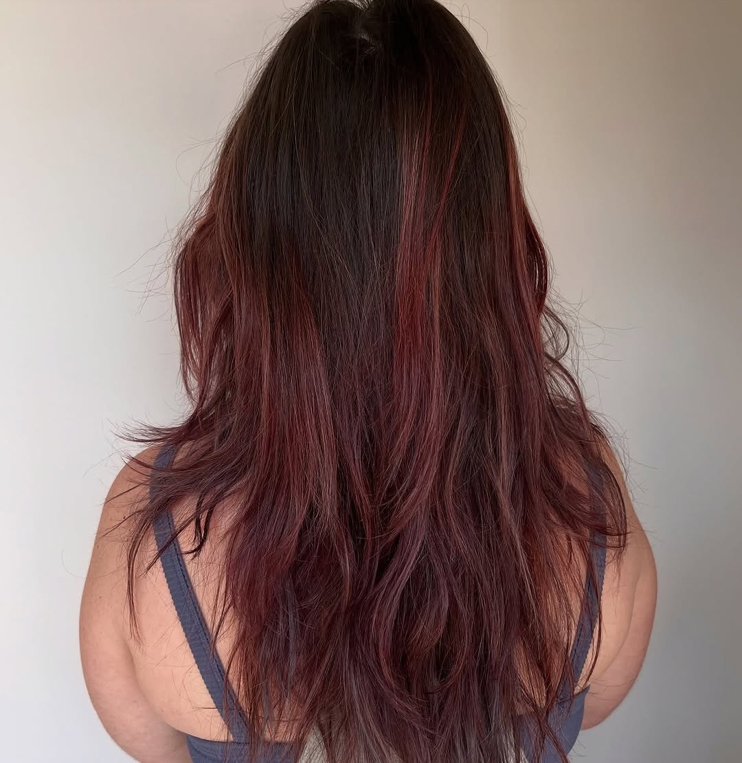 A client at TEAL Hair and Makeup Studio displays rich, dark brunette hair with subtle burgundy-red highlights, styled in soft, natural waves cascading down the back.