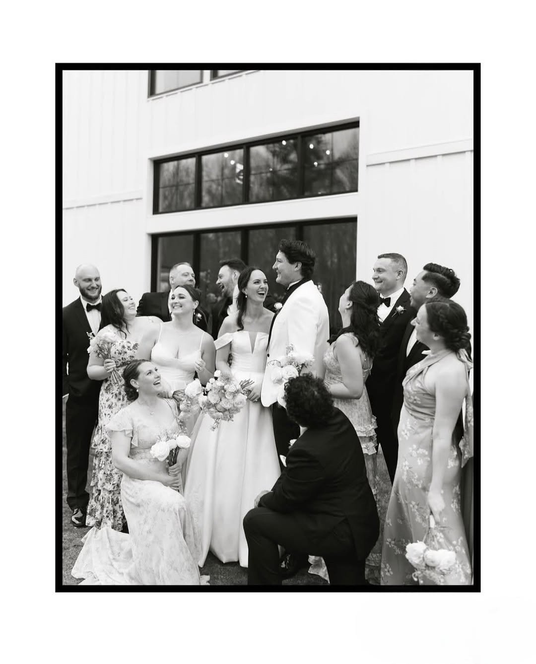 A black-and-white photo captures a joyful wedding party gathered around the bride and groom outside a modern venue, with hair and makeup beautifully styled by TEAL Hair and Makeup Studio.