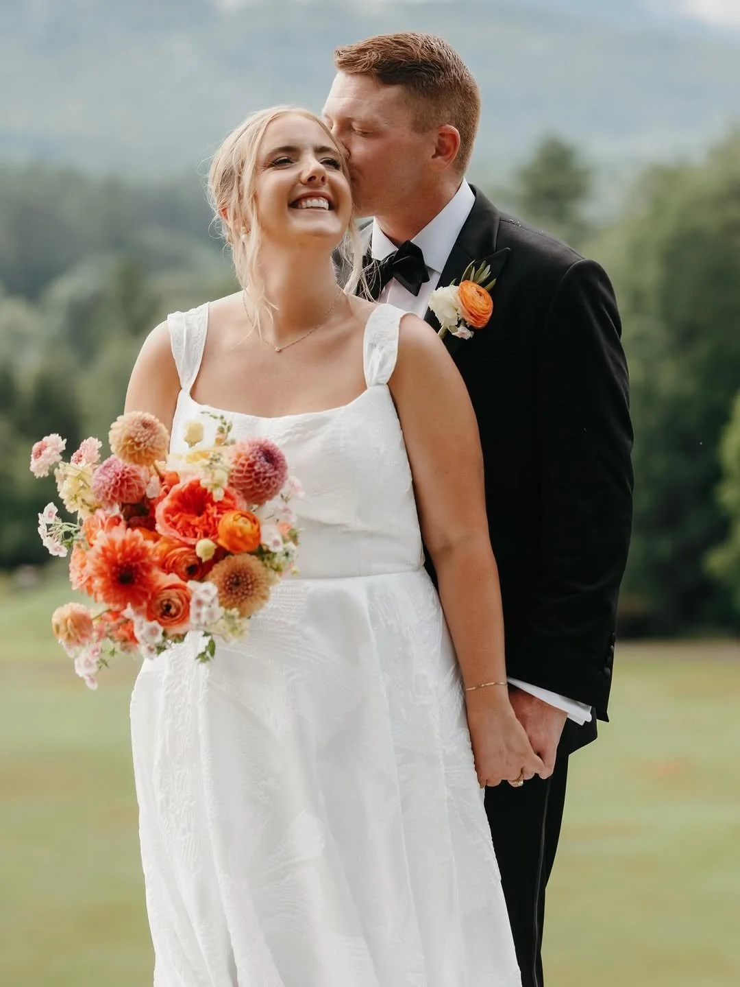  Bride smiling as groom kisses her cheek during an outdoor wedding, showcasing soft, natural bridal hair and makeup by TEAL Hair &amp; Makeup Studio. 