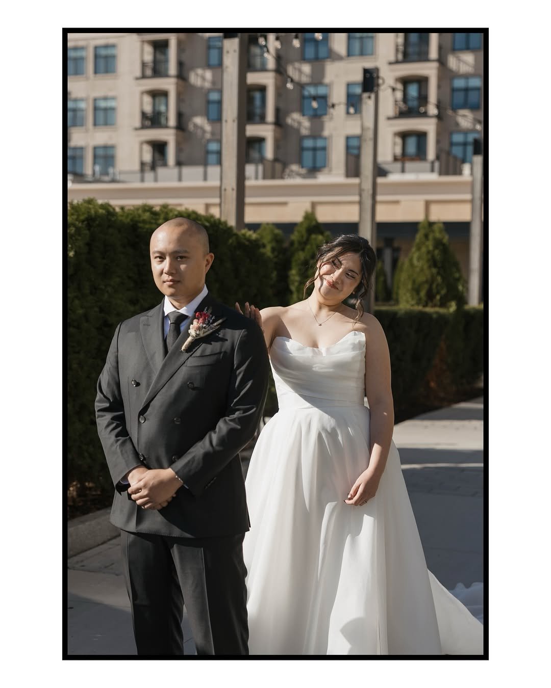  Bride in a strapless white wedding gown standing behind the groom in a black suit during an outdoor wedding, featuring soft bridal hair and makeup by Teal Hair and Makeup Studio. 
