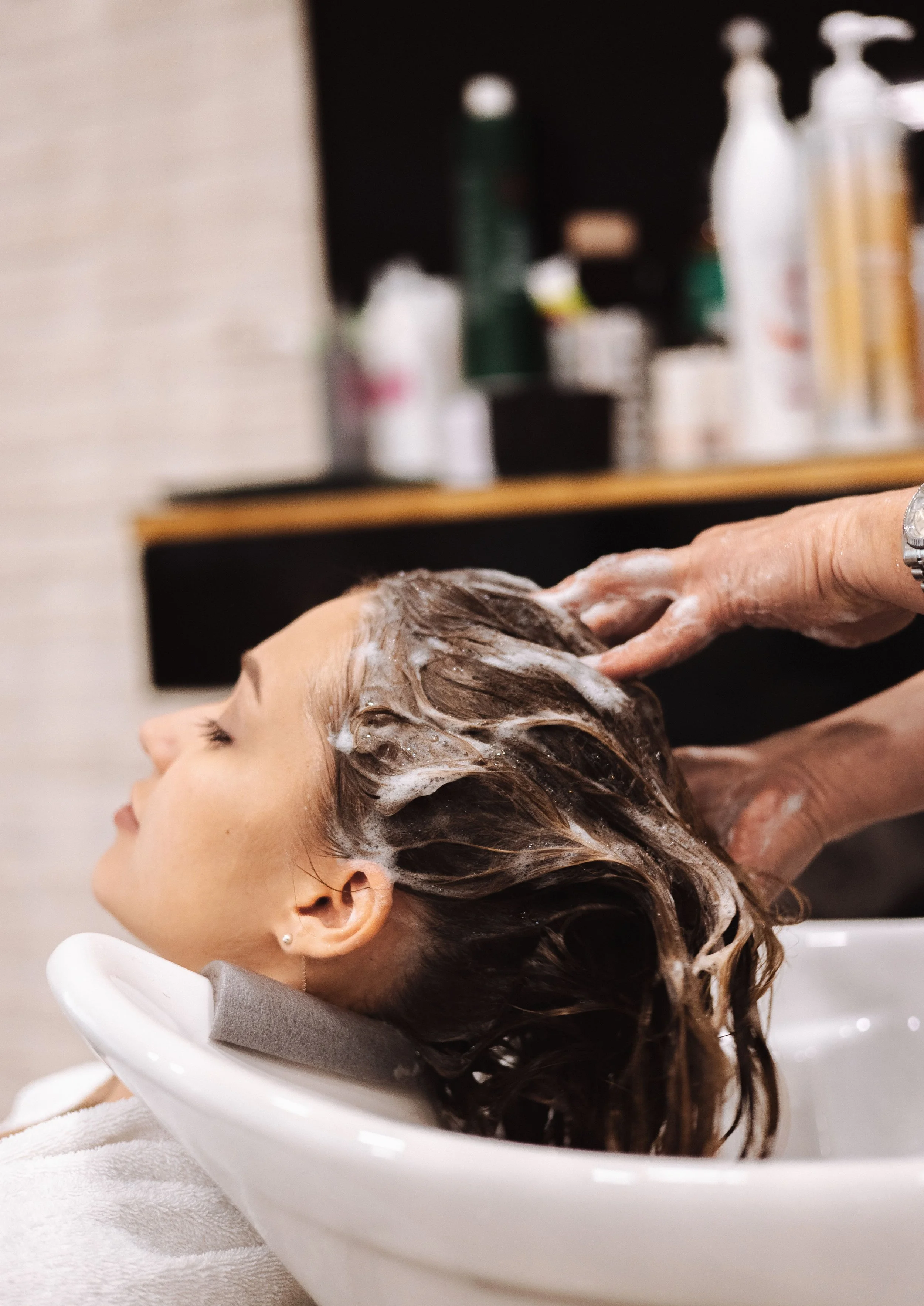 Professional hairstylist gently massaging shampoo into a woman's wet brunette hair at a salon wash station, illustrating a relaxing scalp cleansing and hair care treatment featured by TEAL Hair & Makeup Studio.
