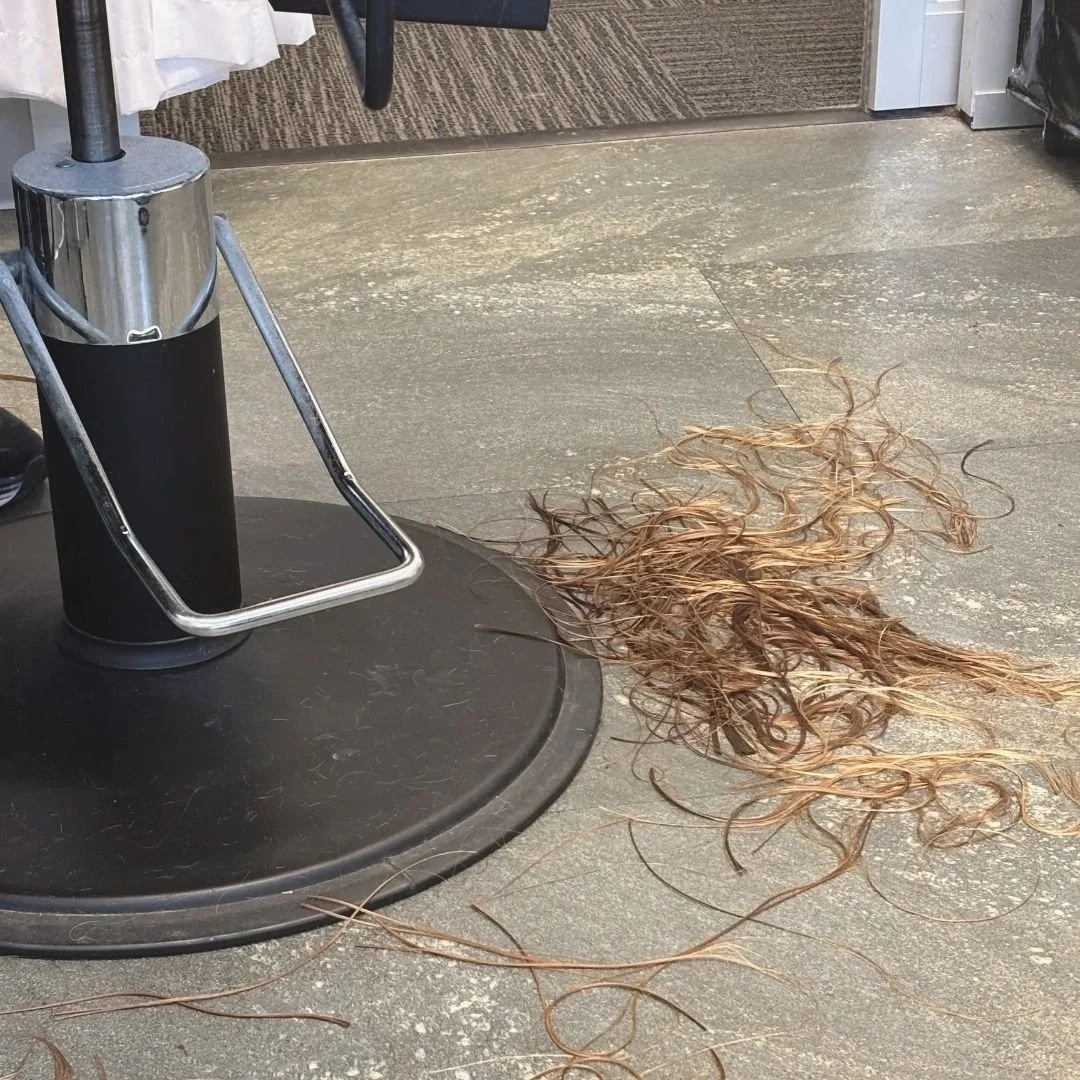 A pile of freshly cut blonde hair rests on a salon floor beside the base of a styling chair.