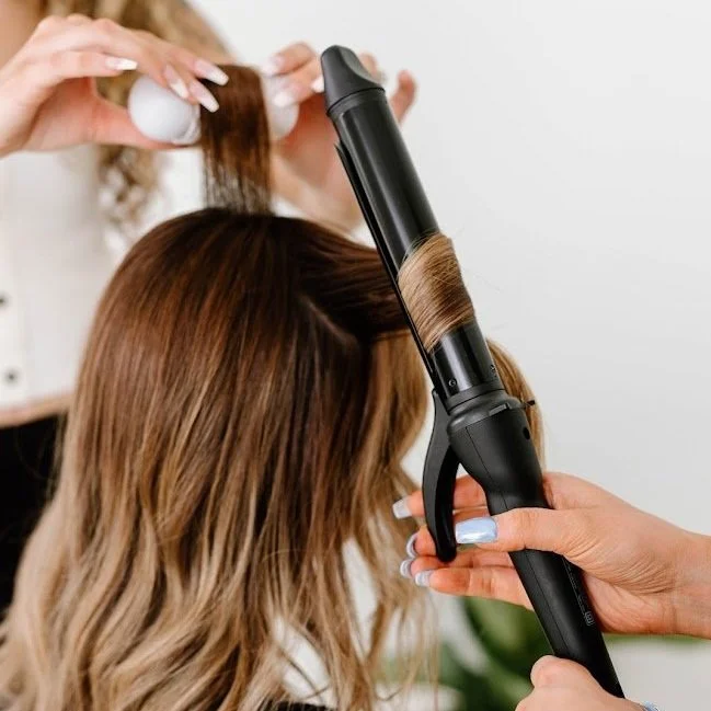 Stylist using a curling iron to create waves in long highlighted hair during thermal styling.