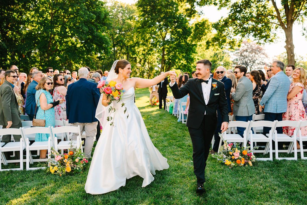 Bride and groom holding hands as they walk down the aisle after an outdoor wedding ceremony, surrounded by cheering guests, colorful floral arrangements, and lush greenery.