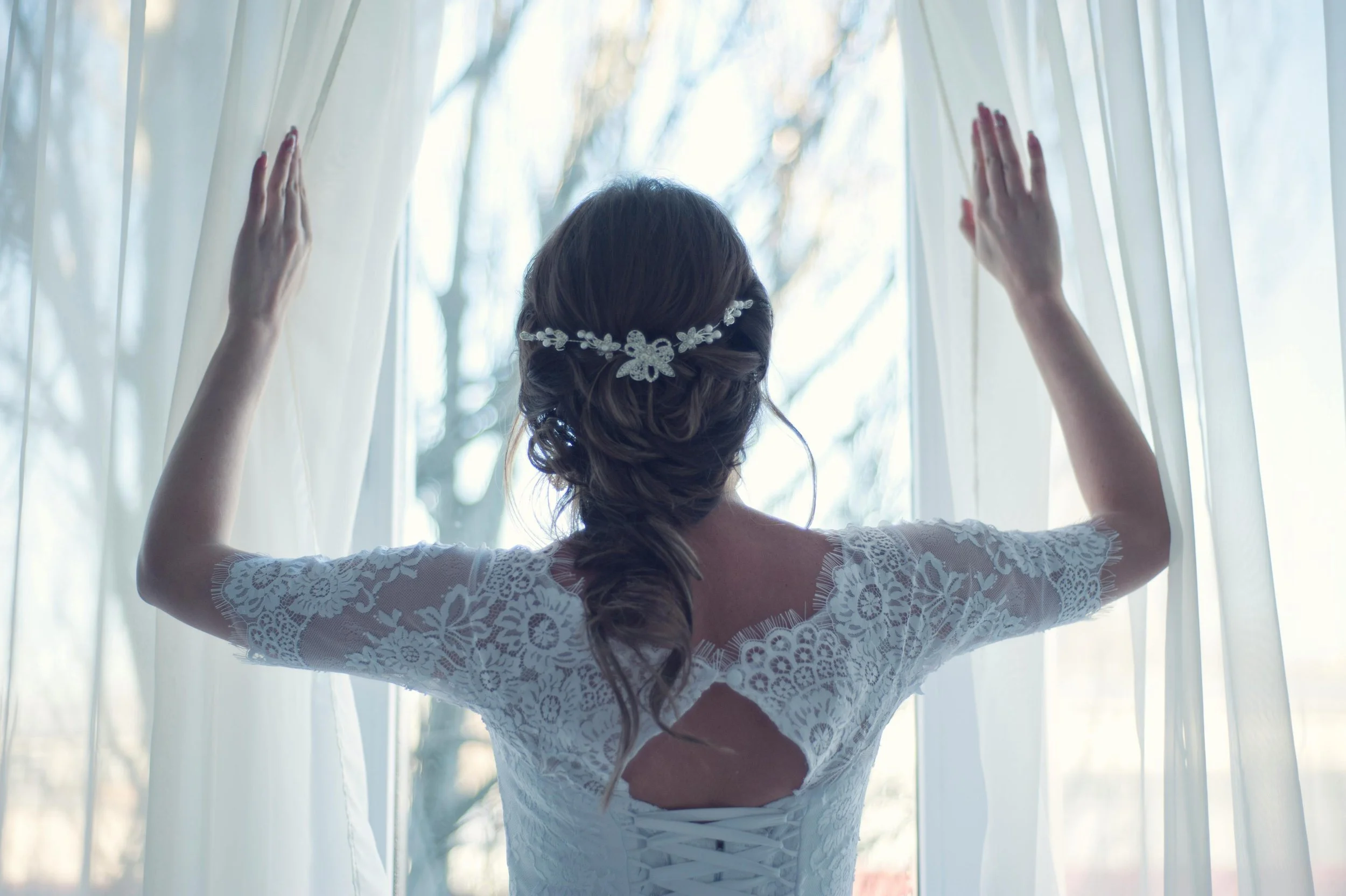 Bride with an elegant braided updo and floral hairpiece in a lace wedding dress opening sheer curtains in soft morning light.