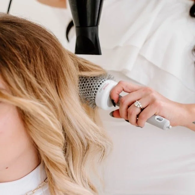 Stylist blow-drying long blonde hair with a round brush during an extended hair styling service.