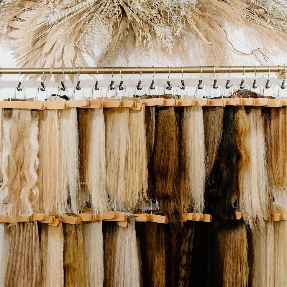 Hair extension wefts in blonde, brunette, and black shades displayed on wooden hangers at Teal Studio