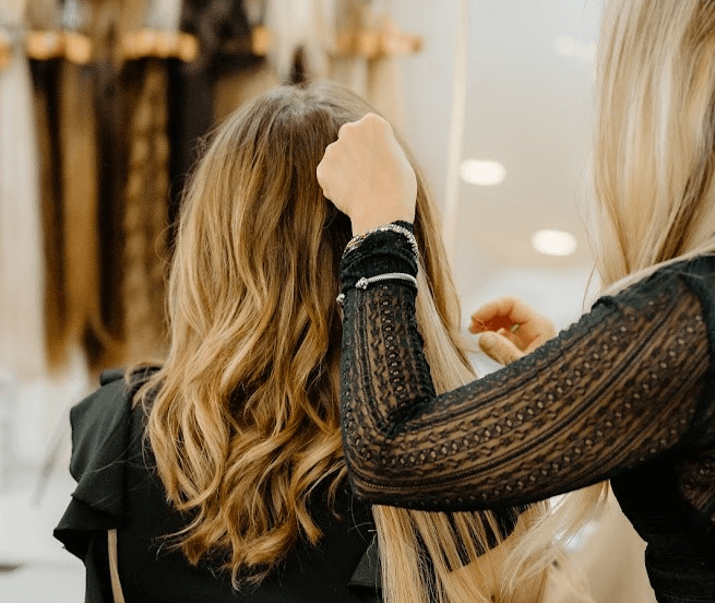 A hairstylist blends long, wavy hair extensions into a client’s natural hair inside a salon.