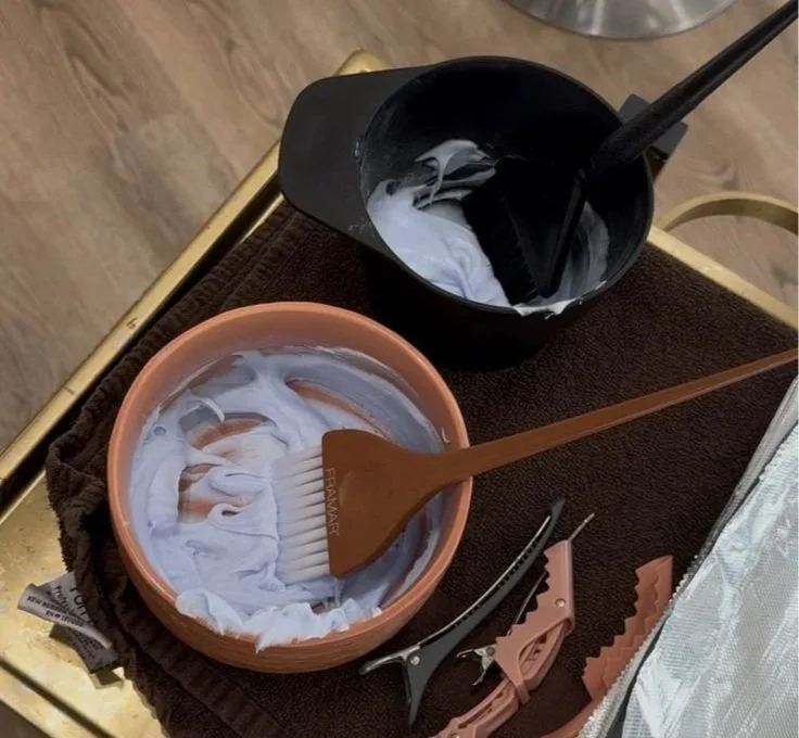 Hair coloring bowls filled with lightening product sit on a salon tray alongside a tint brush and sectioning clips.