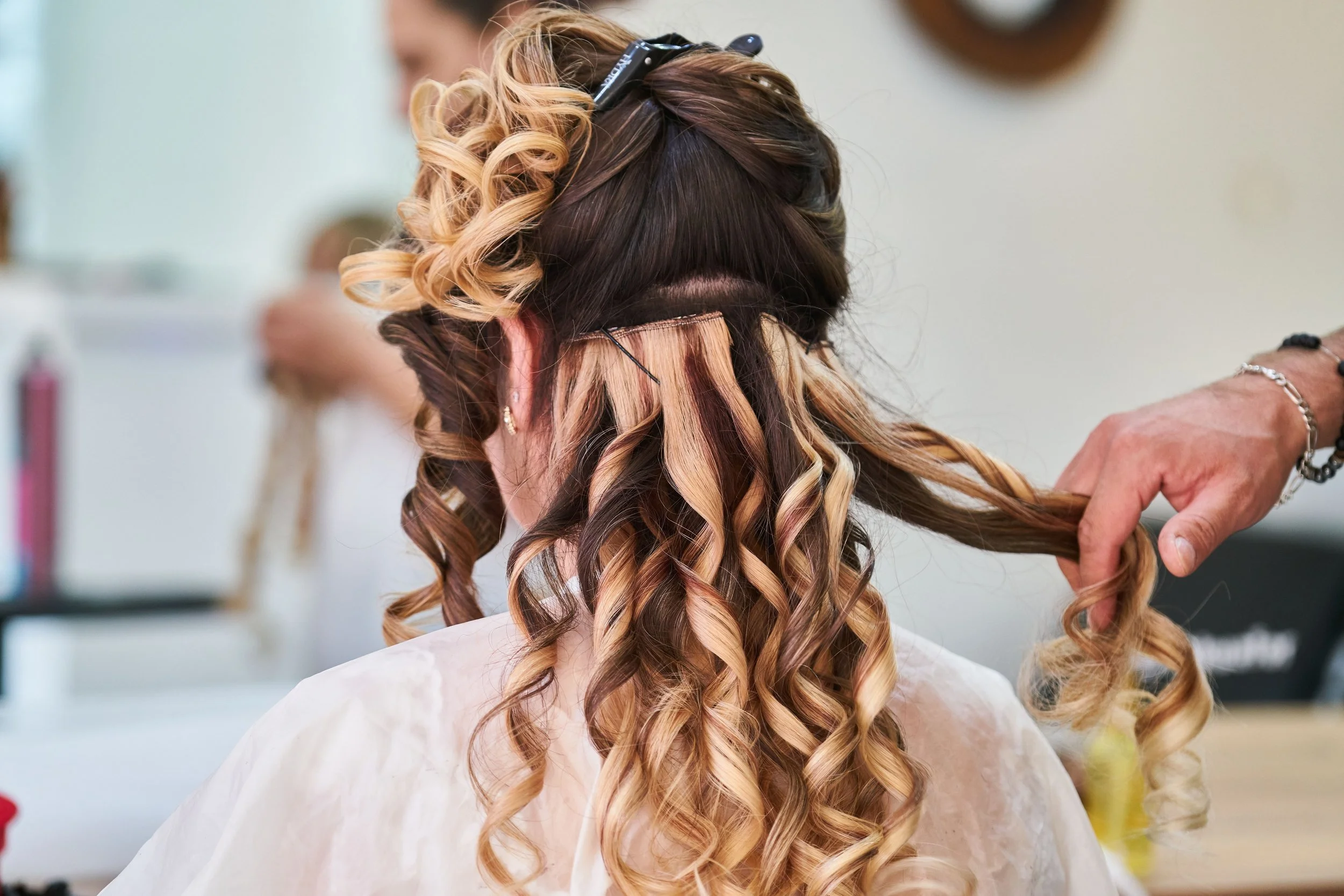 Hairstylist installing and curling blonde hair extensions into a client’s long brunette hair at a salon.