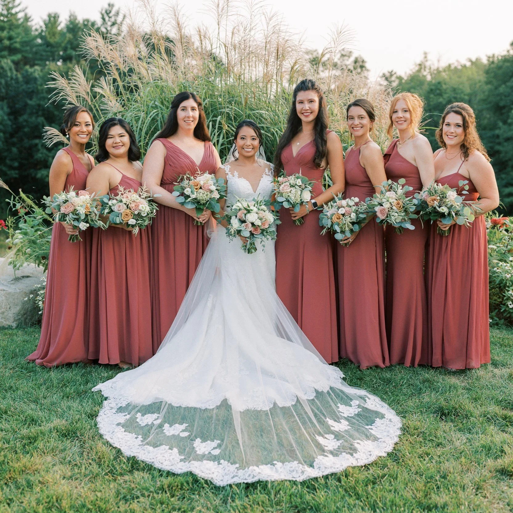 Bride with bridal party in coordinated dresses holding bouquets at an outdoor wedding.